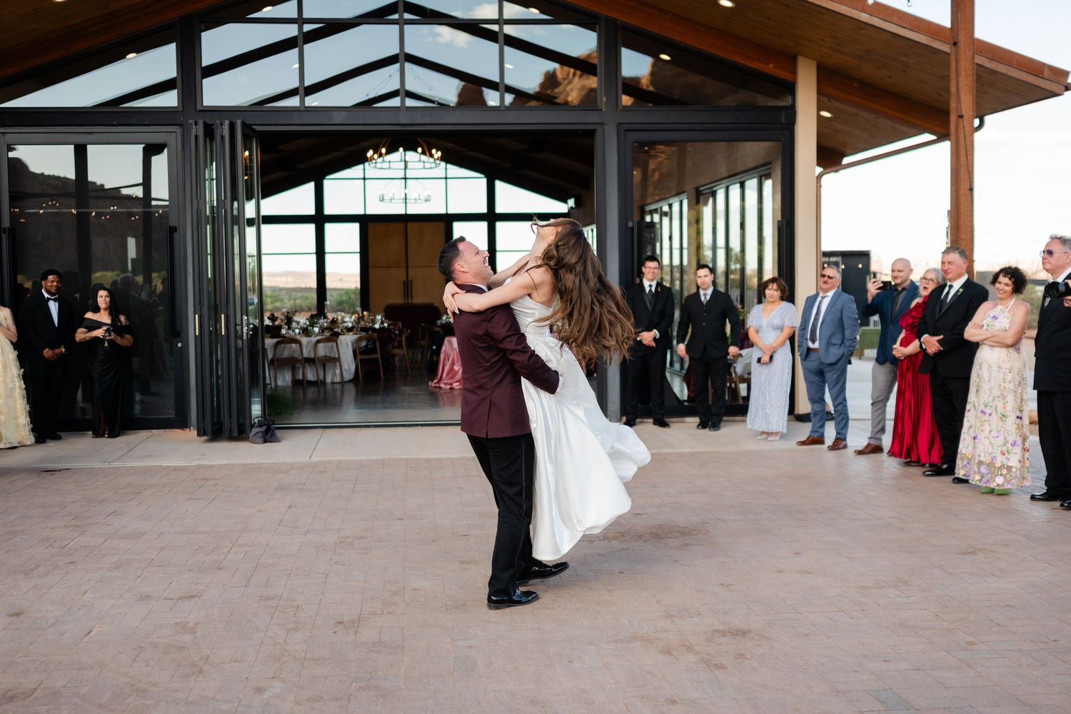 Groom lifts bride during first dance at outdoor wedding venue with guests watching in background.