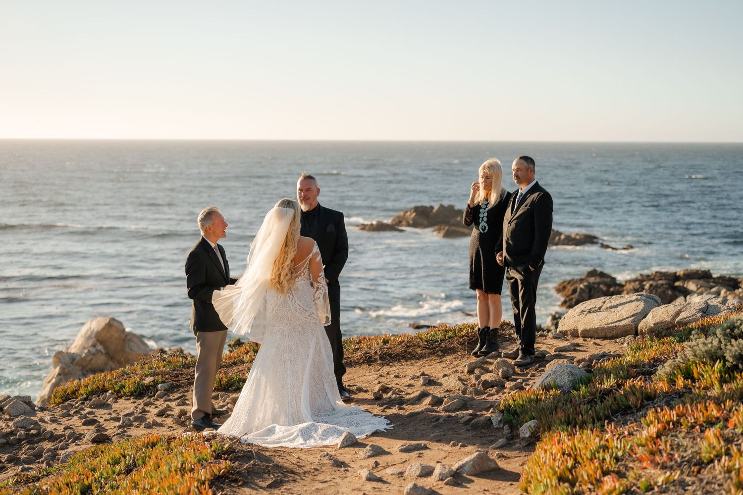 A small group celebrates a wedding ceremony on a scenic cliff overlooking the Pacific Ocean.
