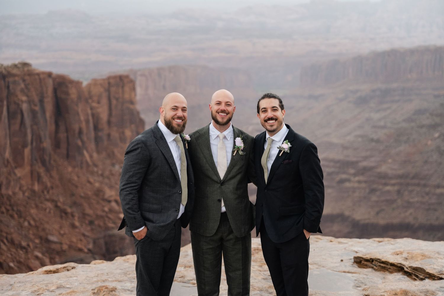 Groomsmen in dark suits pose together on rocky desert overlook with canyon views.