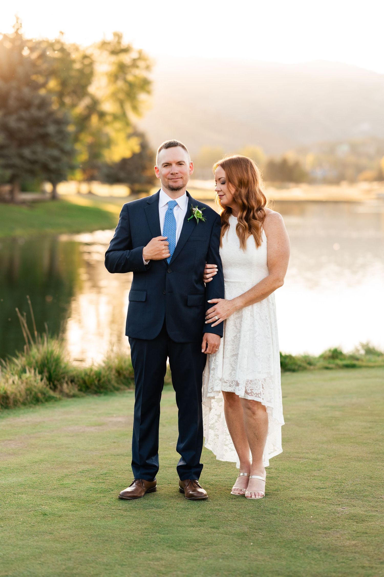 A couple poses together on grassy lawn beside a lake during sunset.