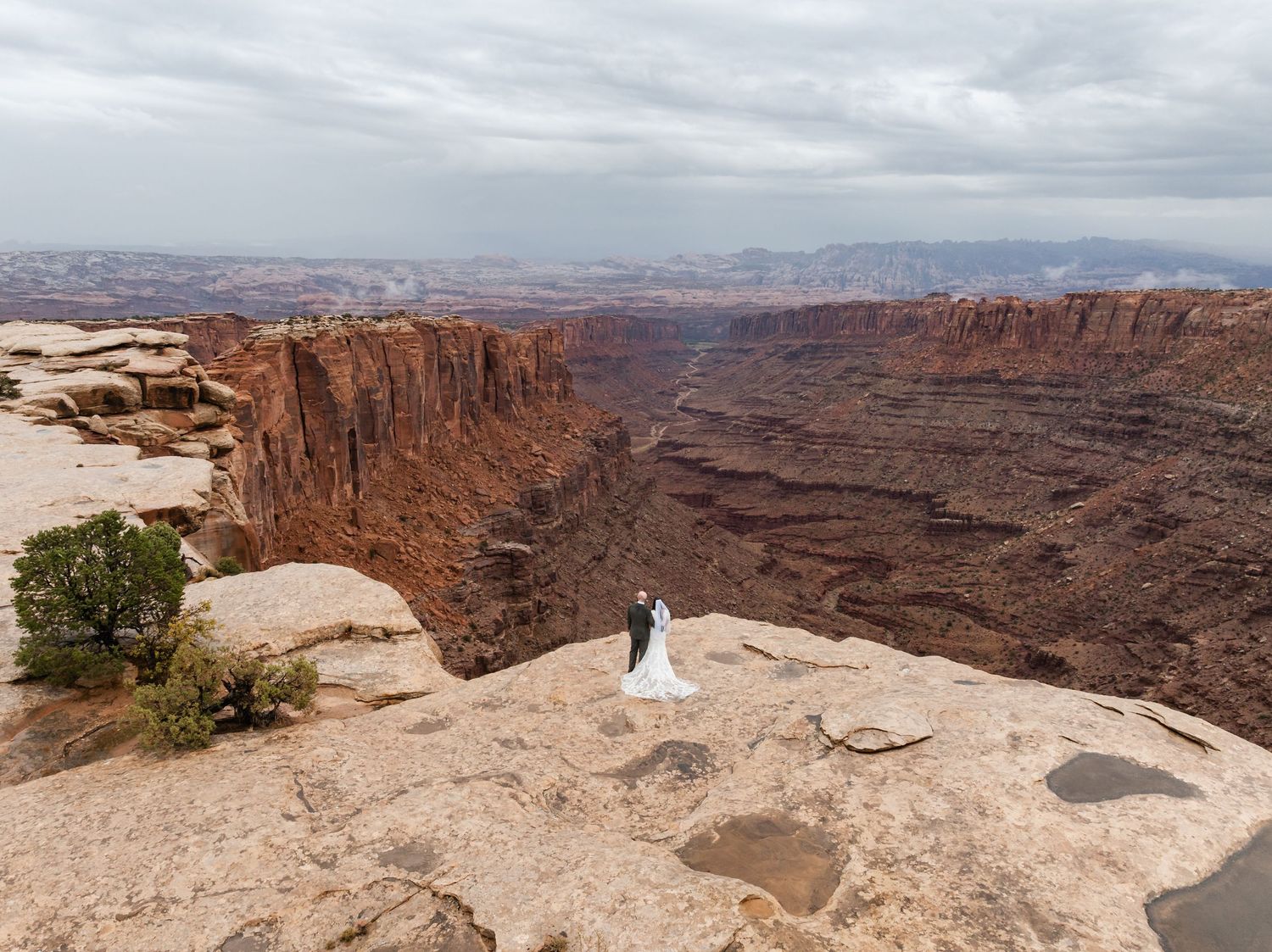 A couple in wedding attire stands at the edge of a dramatic canyon overlook at Canyonlands National Park in Utah.