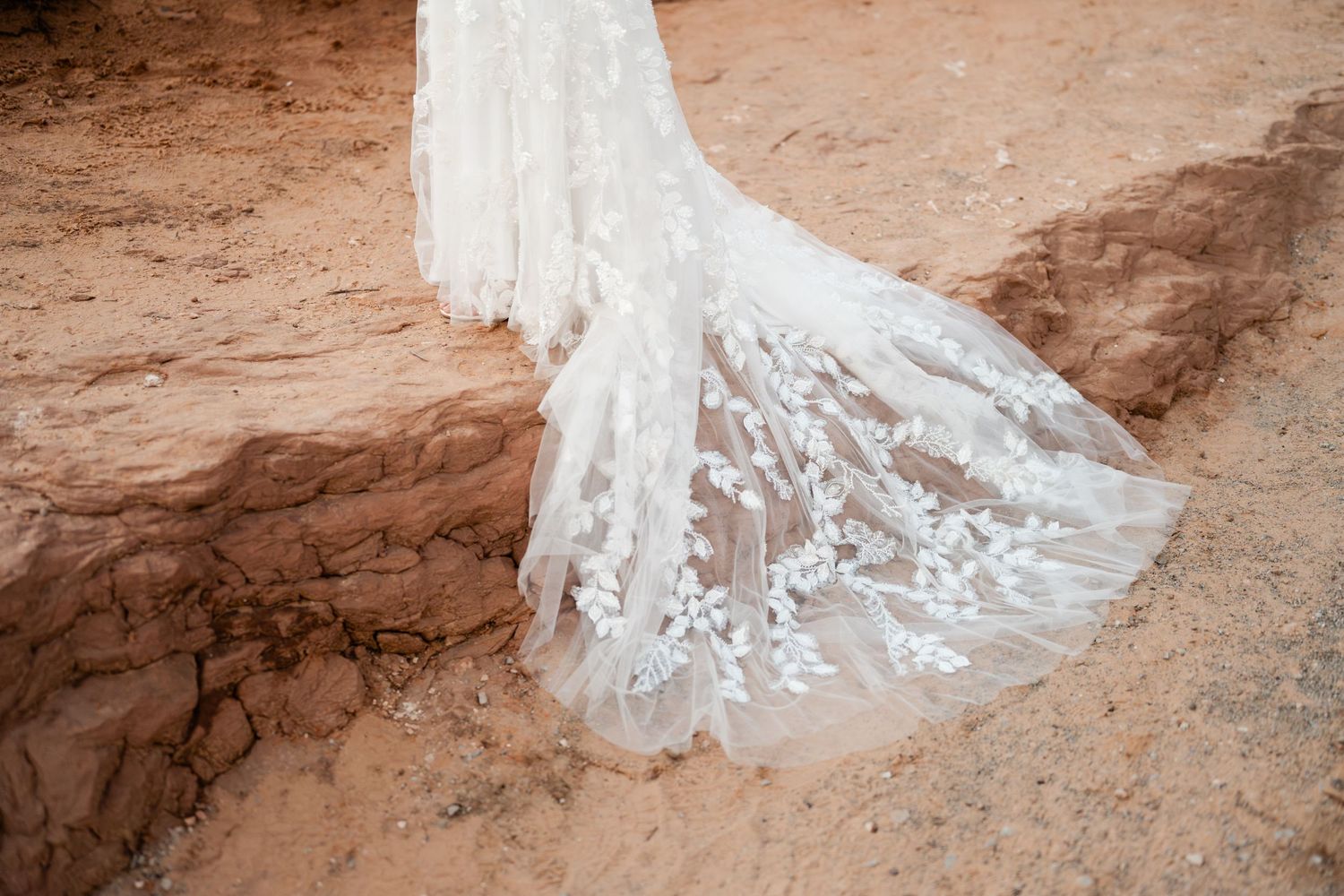 A detailed view of an intricate lace wedding dress train against red desert sand.