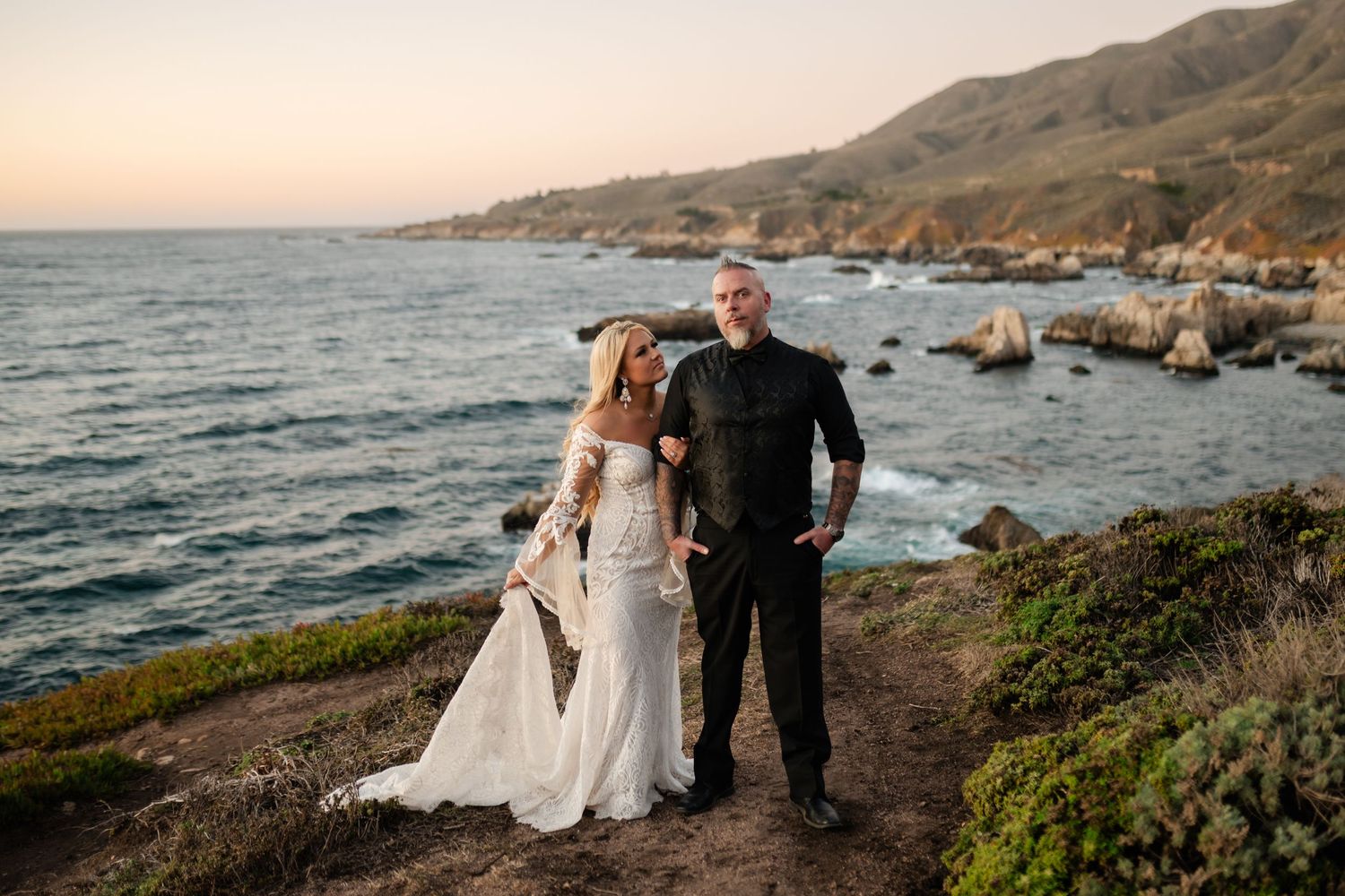 A couple in wedding attire standing on coastal cliffs at sunset.