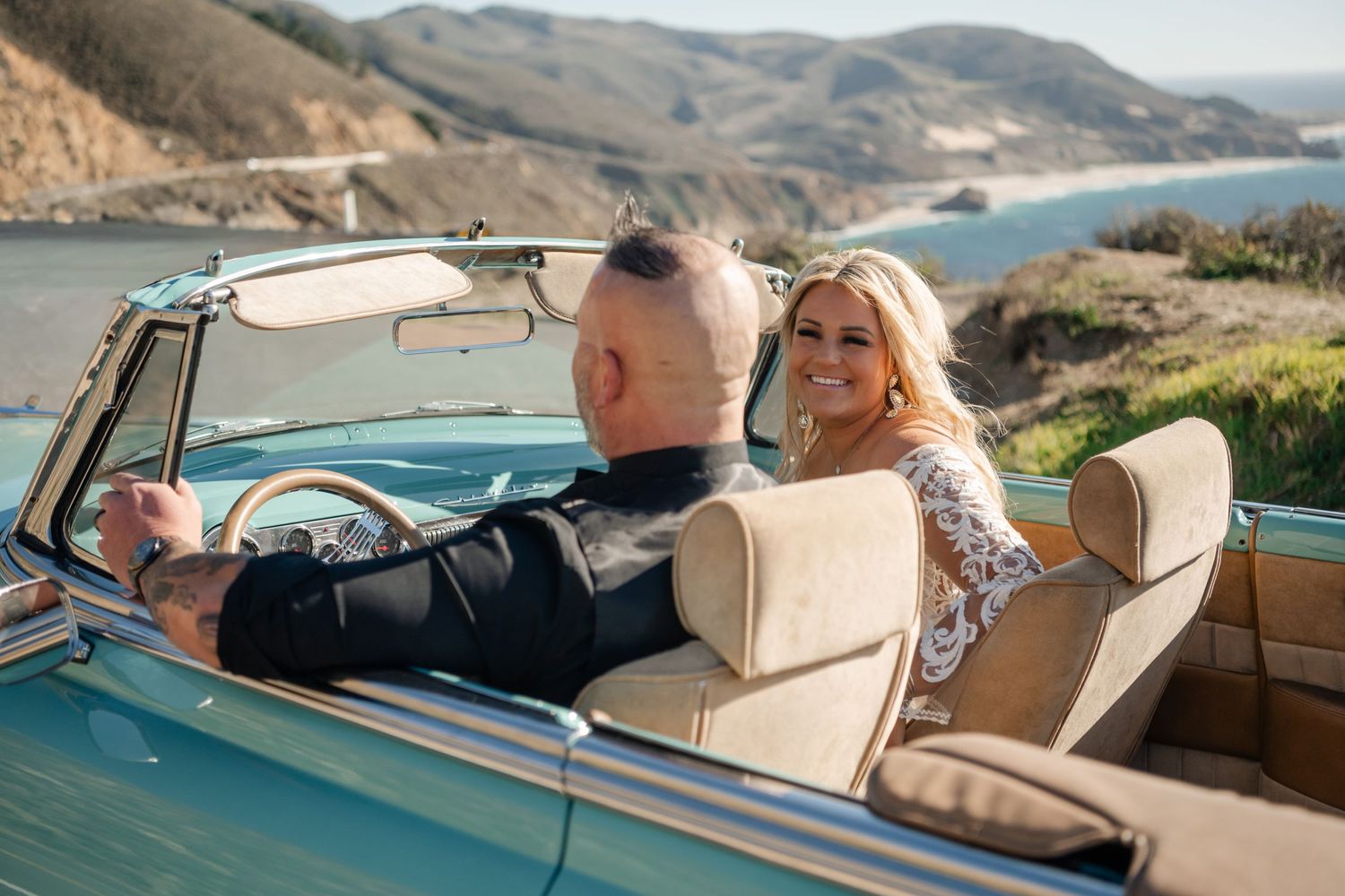 A classic turquoise convertible car drives along a scenic coastal mountain road.