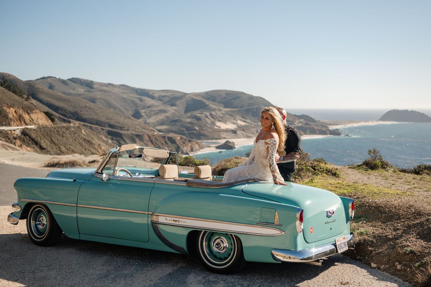 A vintage turquoise convertible car parked along a scenic coastal cliff overlooking the ocean at sunset.