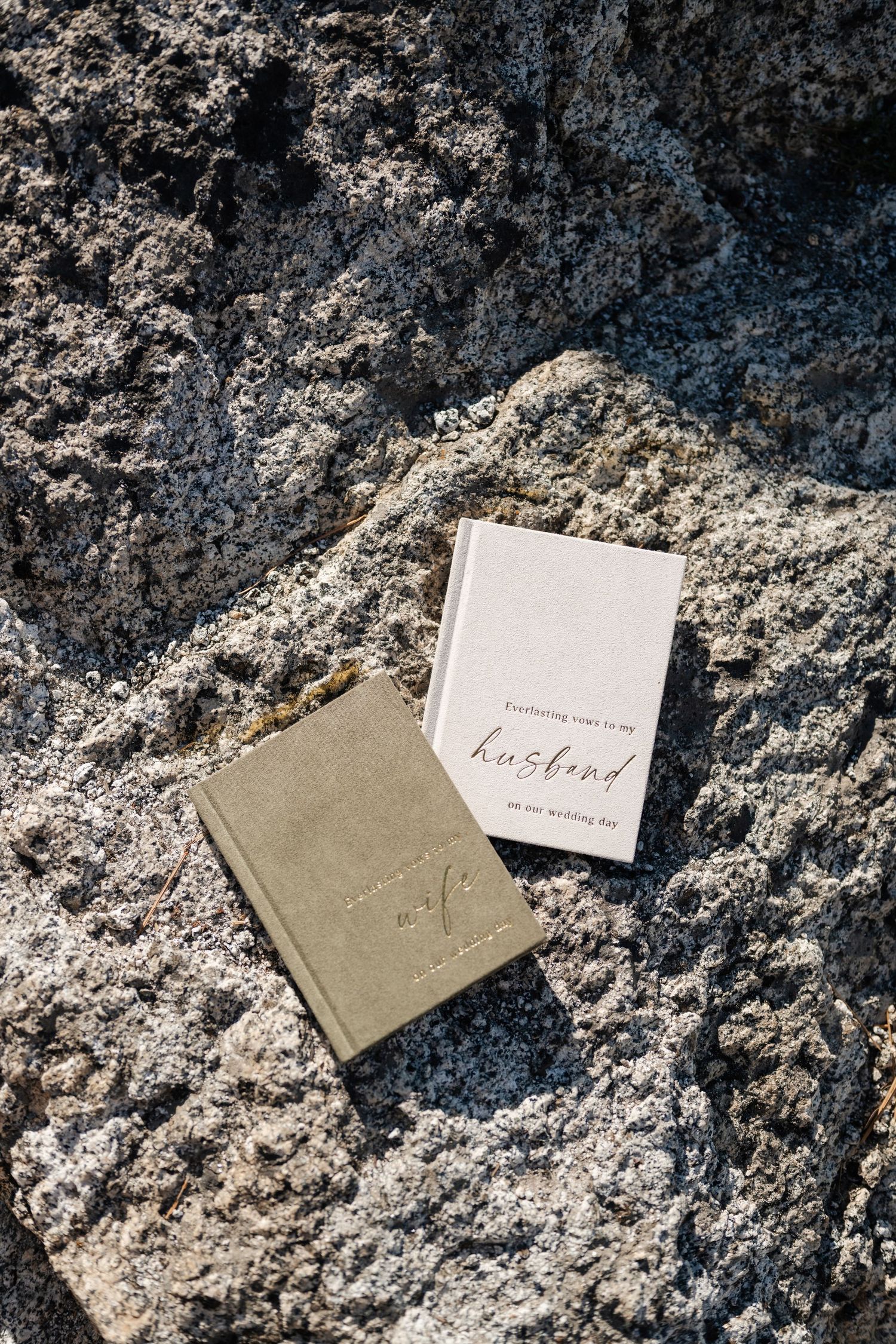 Two wedding vow books resting on textured rock, one olive green and one cream colored reading husband on wedding day.