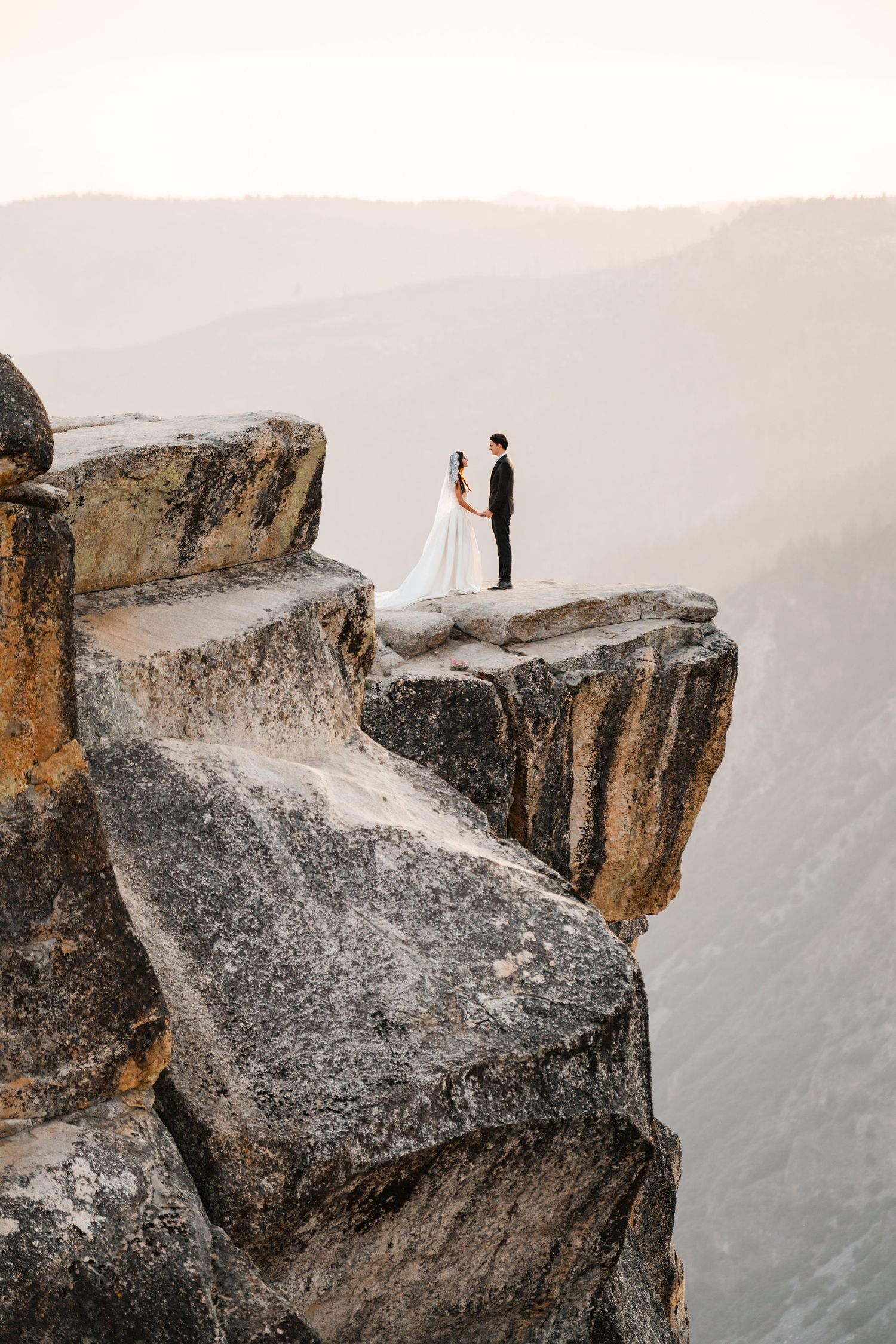 Bride and groom stand atop a dramatic rocky cliff at sunset with misty mountain valleys below.
