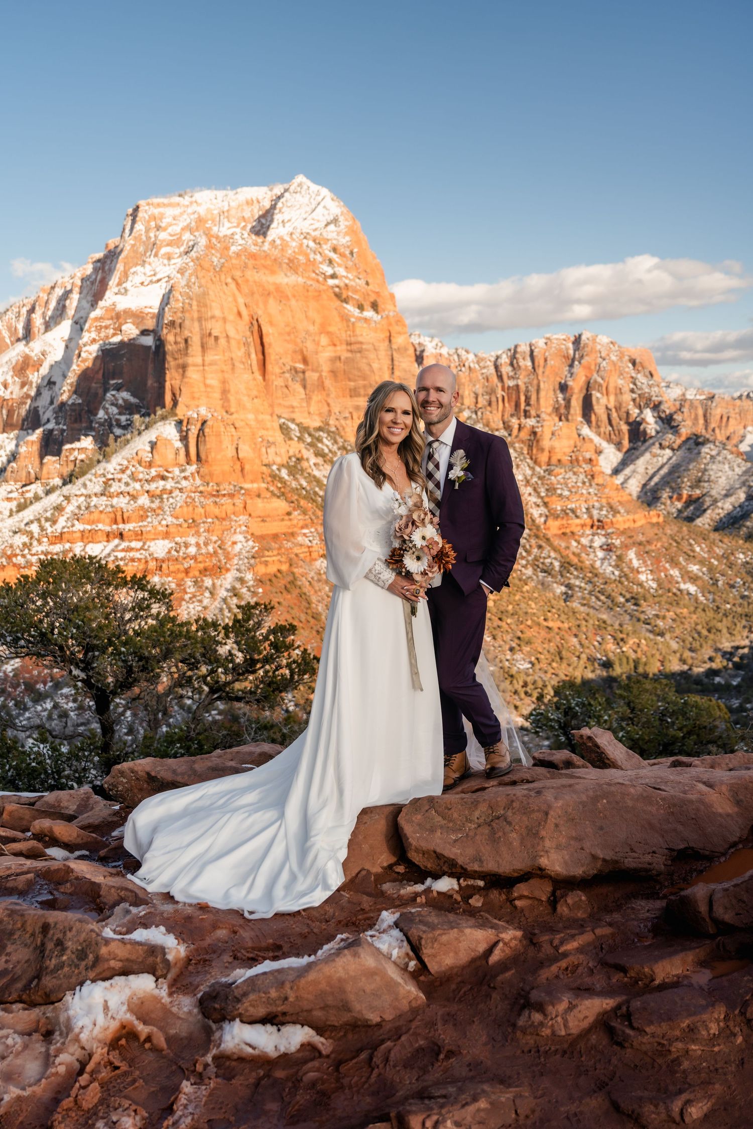 A couple in wedding attire poses against stunning red rock formations and mountain vistas at sunset in Zion National Park.