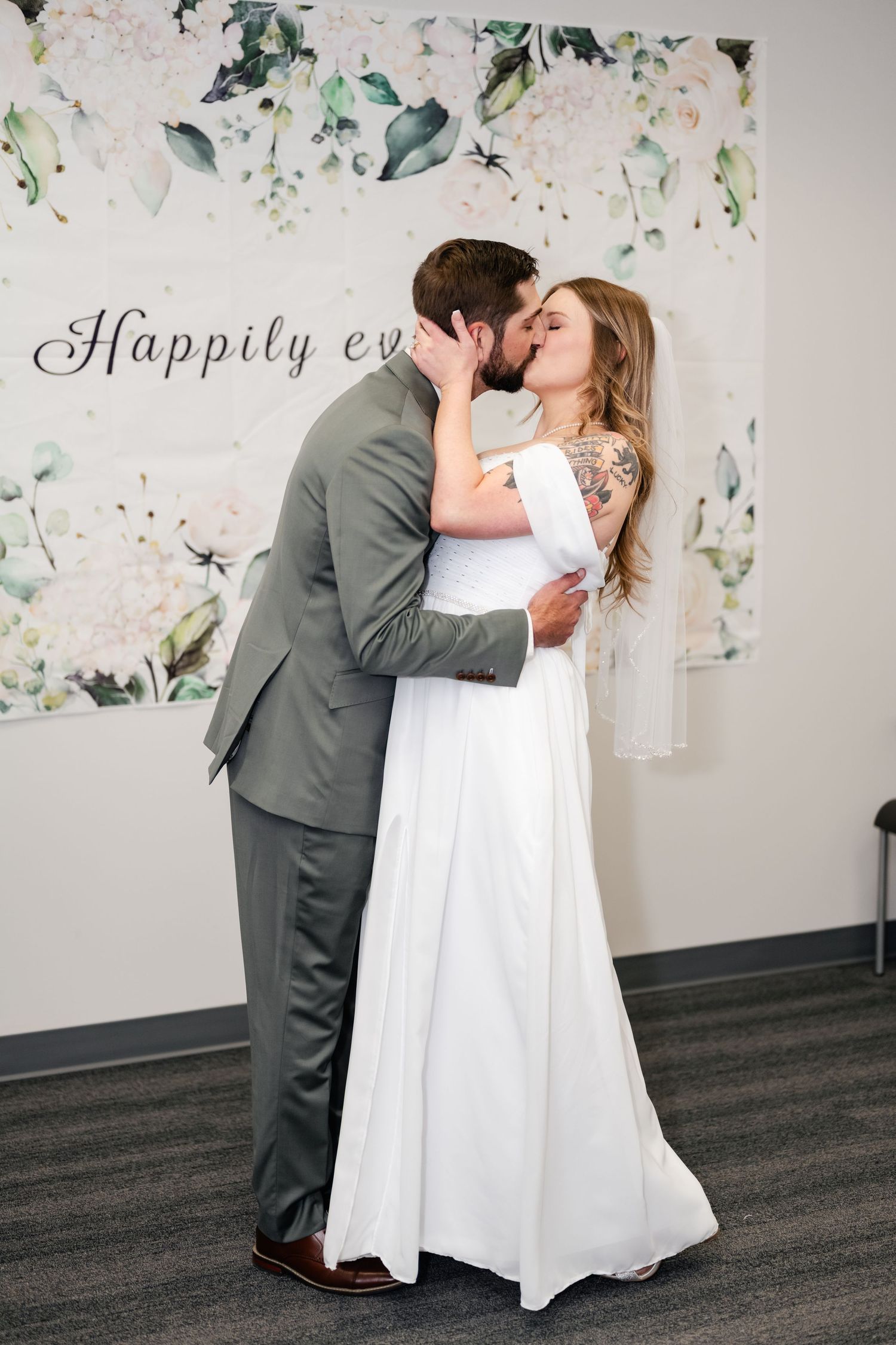 A couple shares a romantic kiss in front of a floral backdrop with 'Happily ever' text during their wedding celebration.