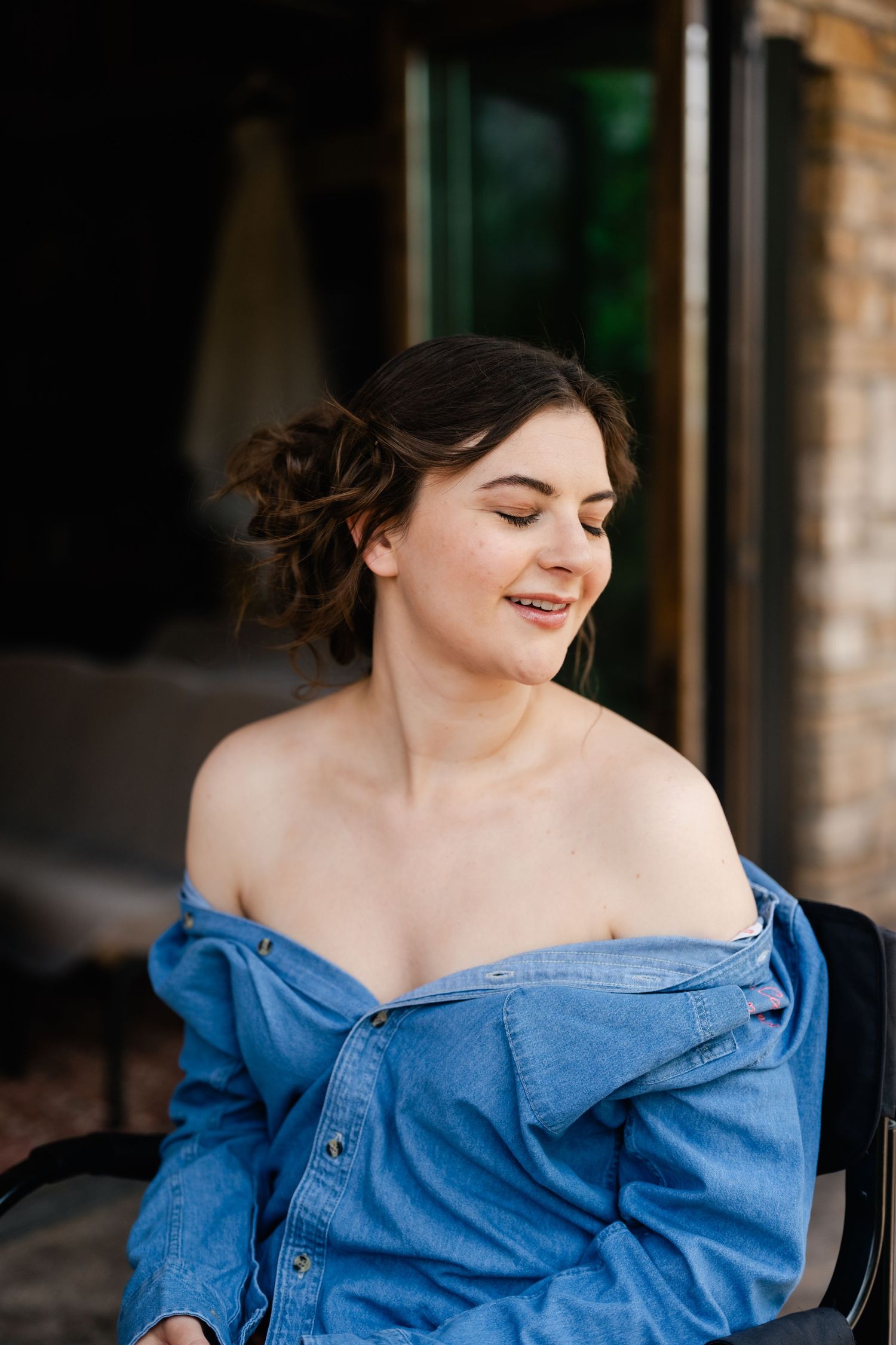 Person in off-shoulder denim jacket smiling with eyes closed and styled updo hairstyle.