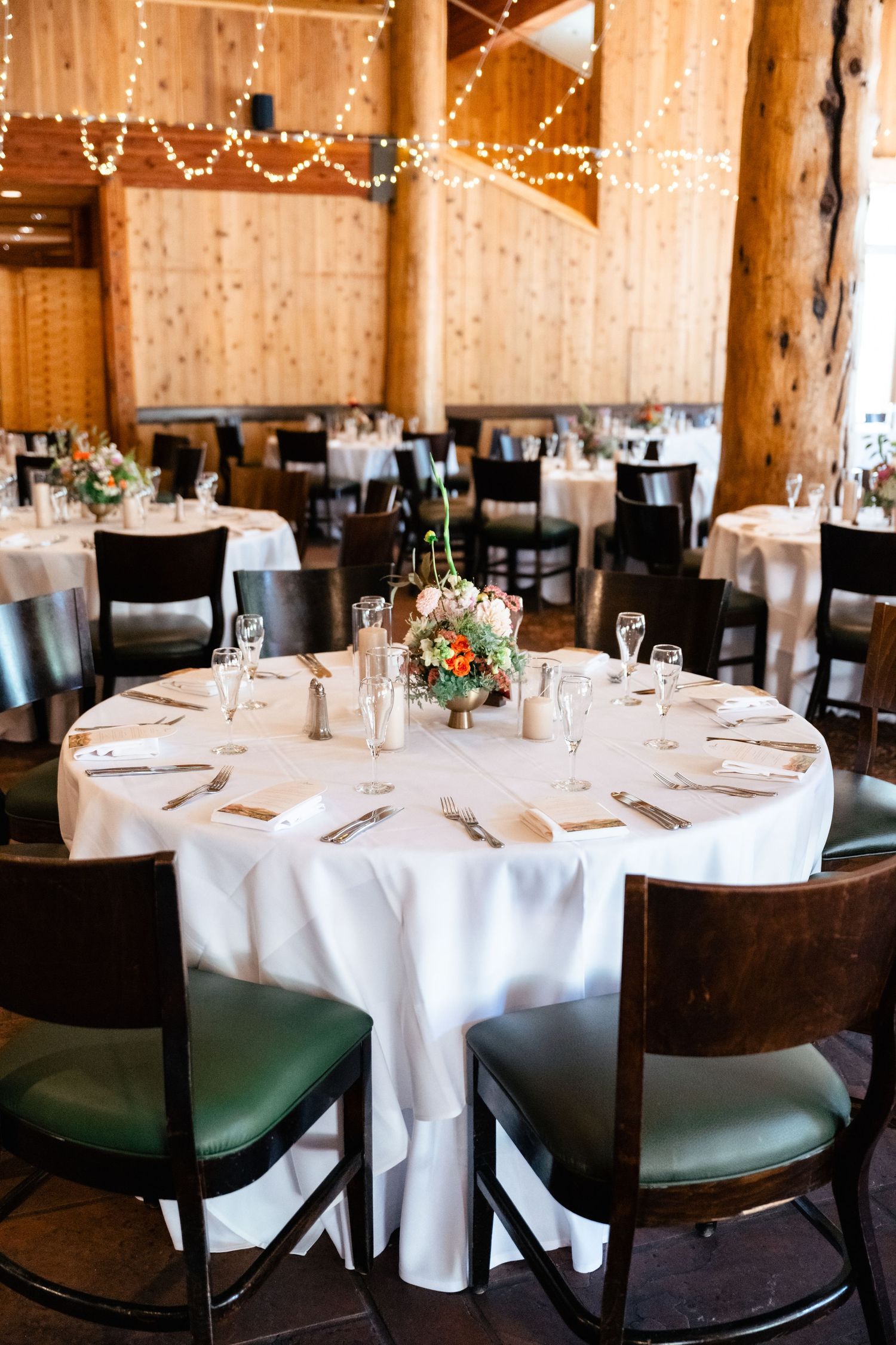 Rustic wedding reception table setting with white linens, dark wooden chairs and twinkle lights in a barn venue.