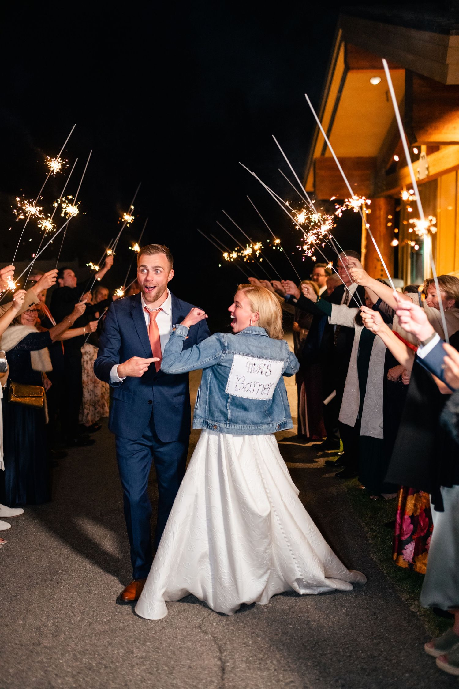 A couple celebrates with sparklers during their wedding exit while the bride wears a customized denim jacket.