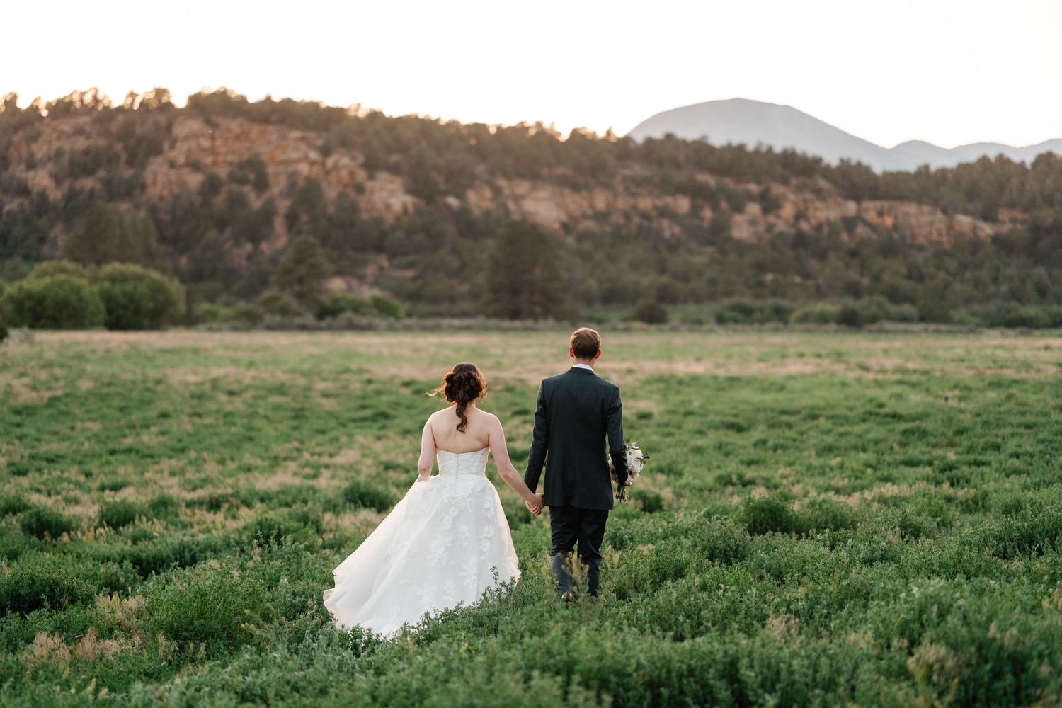 A couple walks hand in hand through a green meadow with mountains in the background.