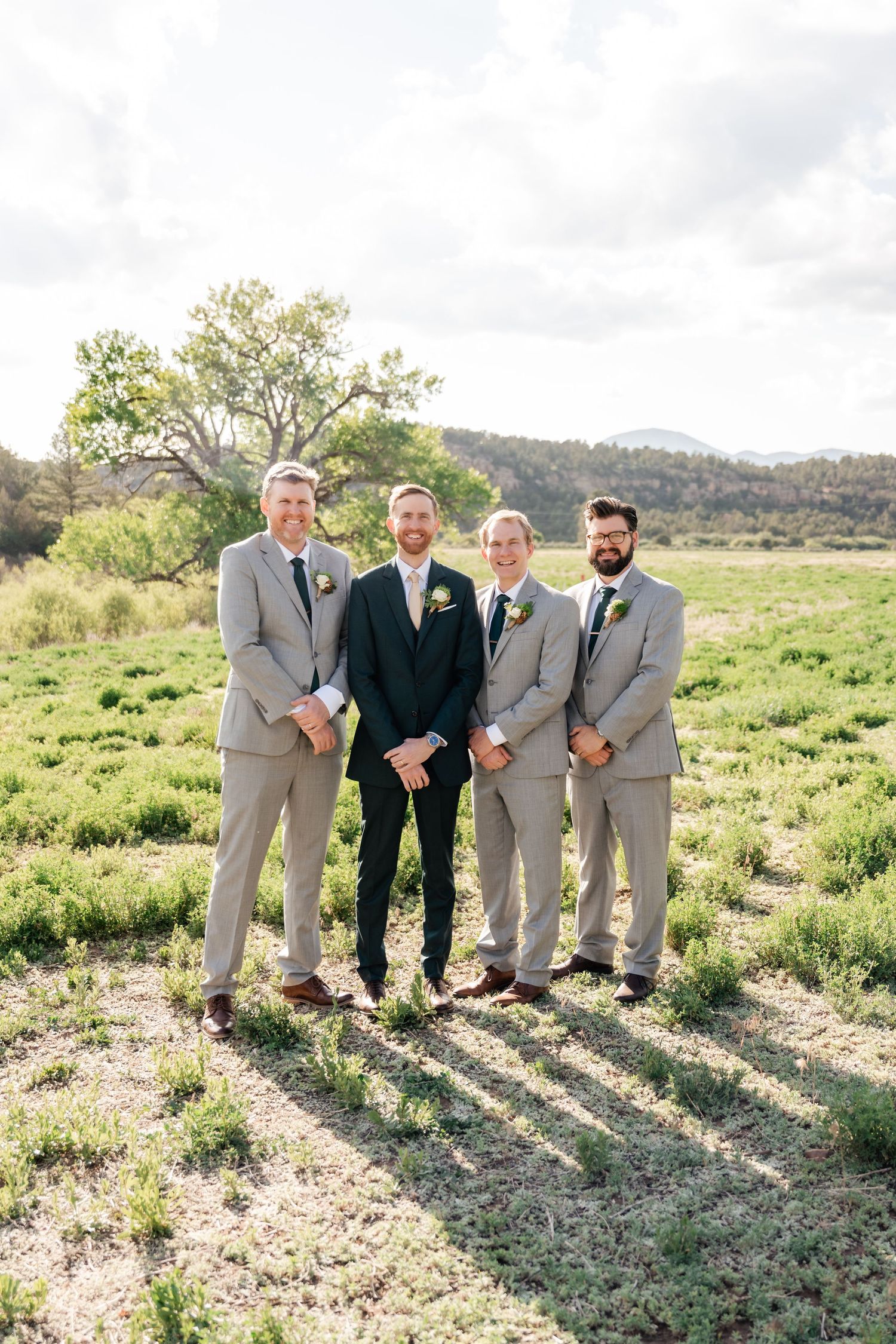 Groomsmen in grey and navy suits stand together in a rustic outdoor setting with hills in the background.