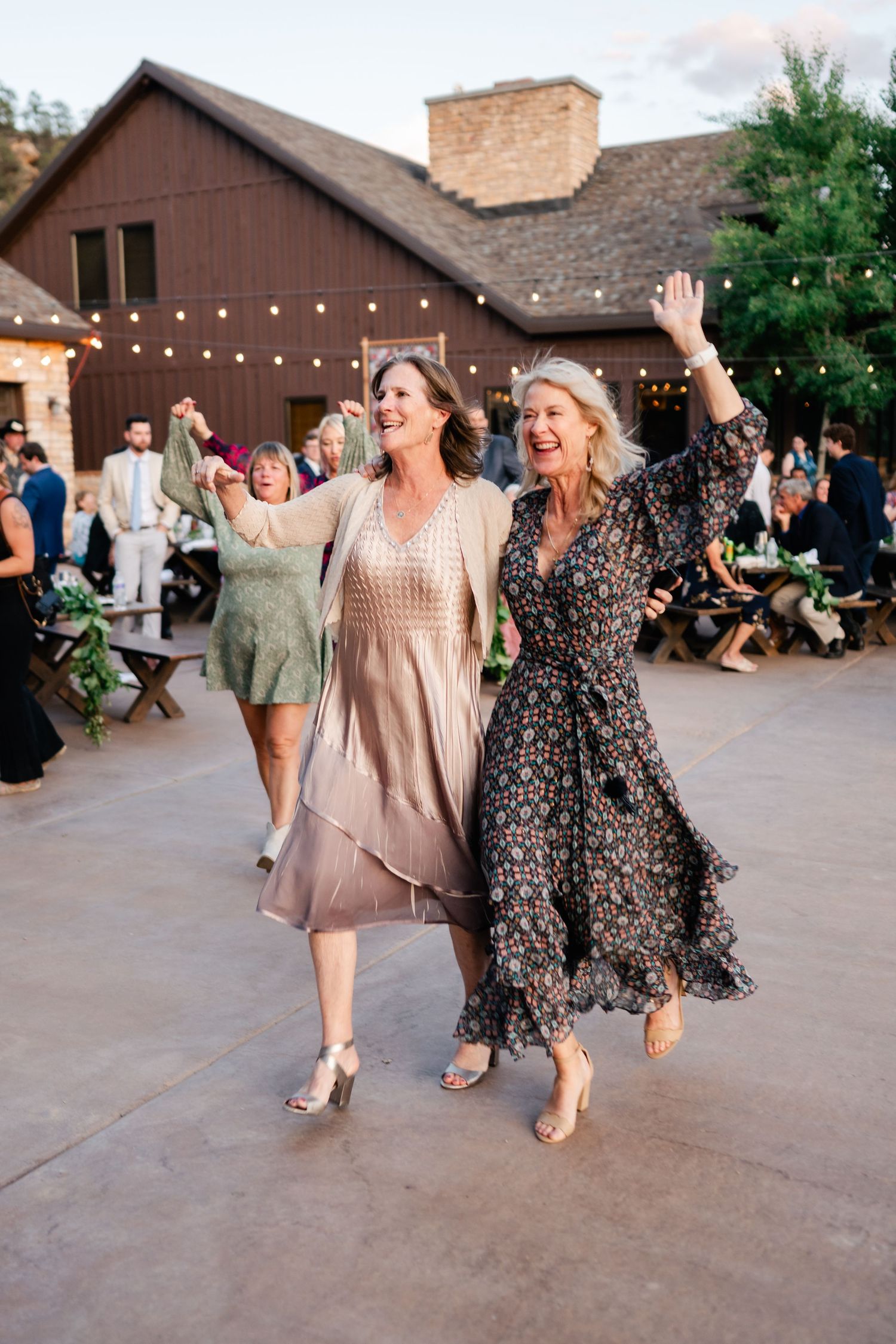 Two women in flowing dresses dance joyfully at an outdoor wedding reception.