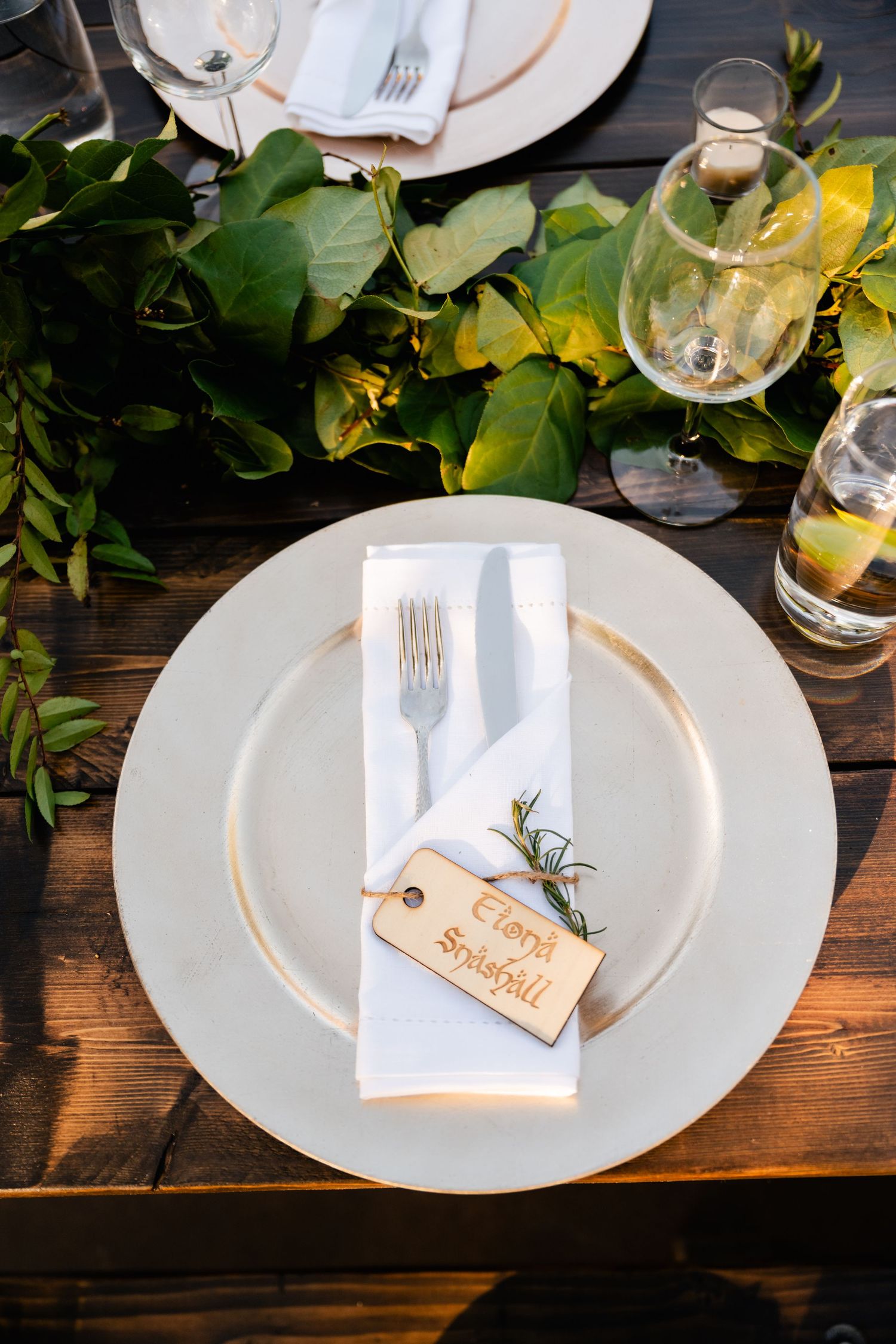 Place settings at a rustic wedding reception feature white plates with name cards and greenery on wooden tables.
