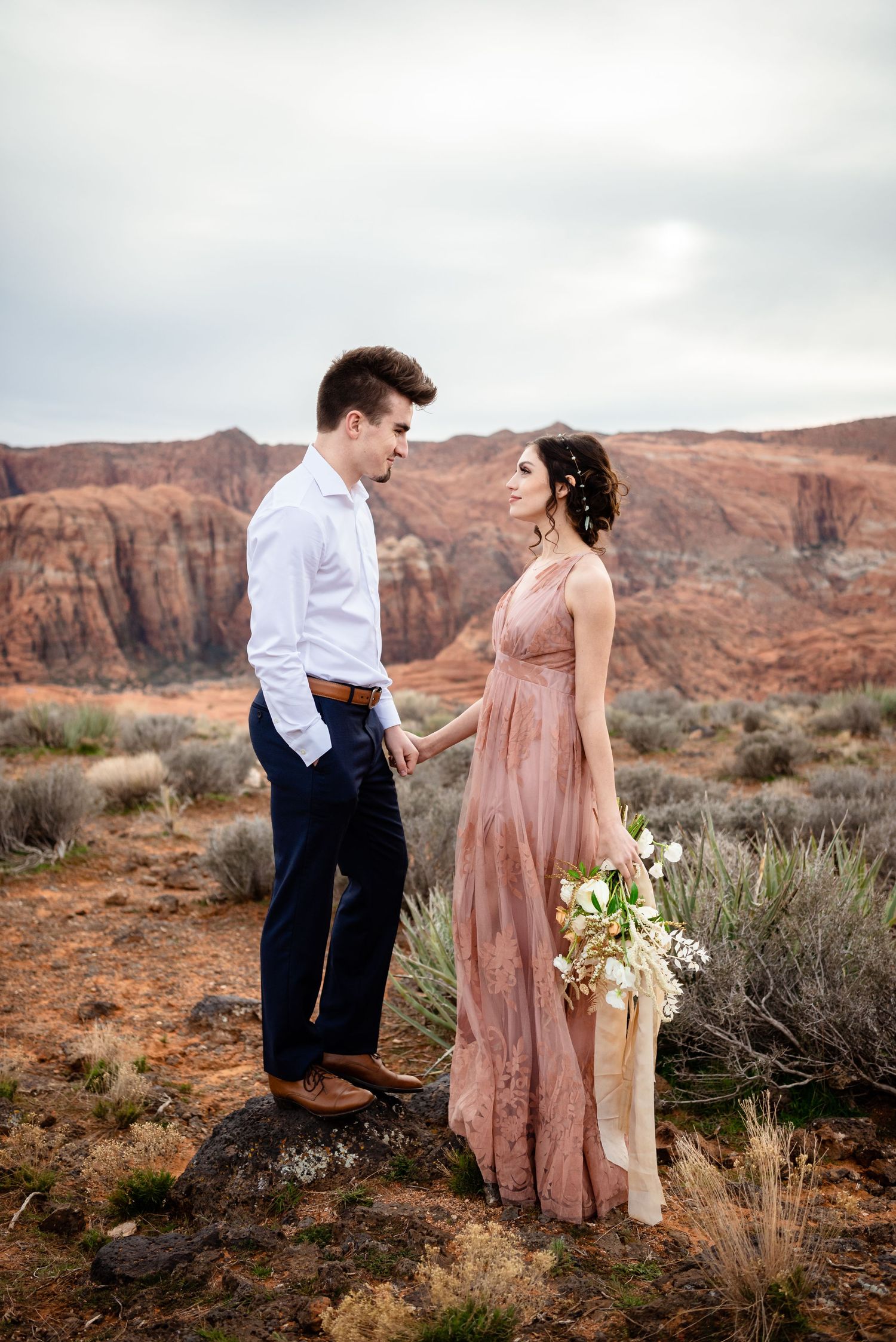 A couple shares a romantic moment against the backdrop of red rock cliffs and desert brush in Utah. Snow Canyon Photographer.