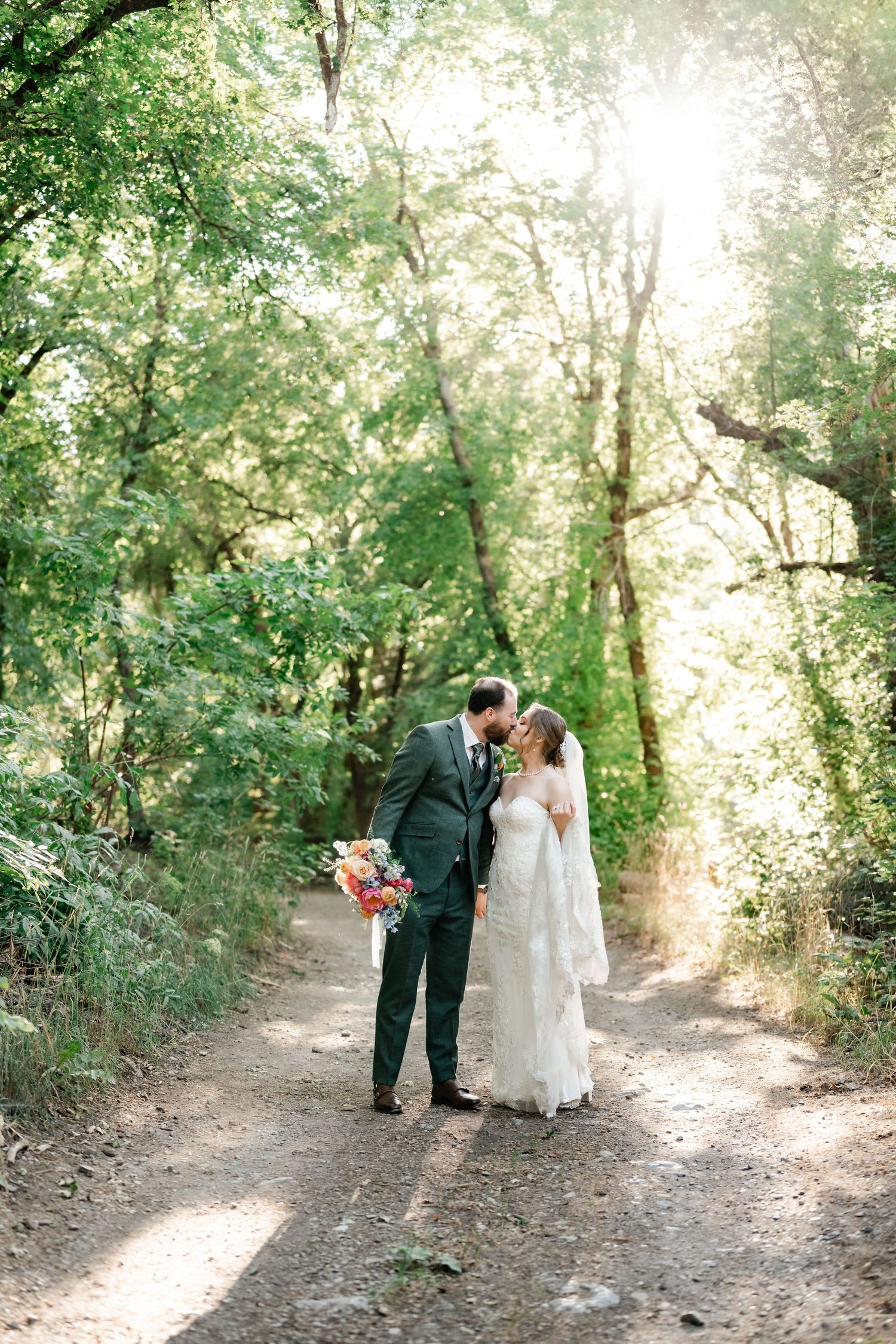 A couple shares an intimate moment on a sunlit forest path surrounded by lush greenery. Millcreek Inn Wedding Photographer.