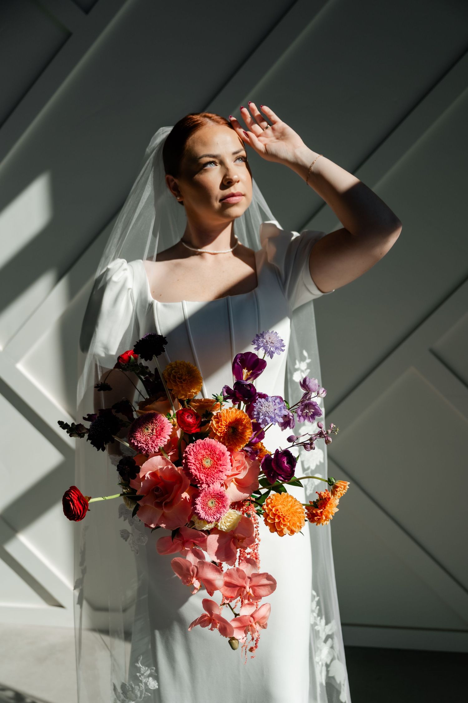 A bride holds a vibrant bouquet of coral, orange and purple flowers against a sunlit wall with diagonal shadows. Enchanted Event Center.