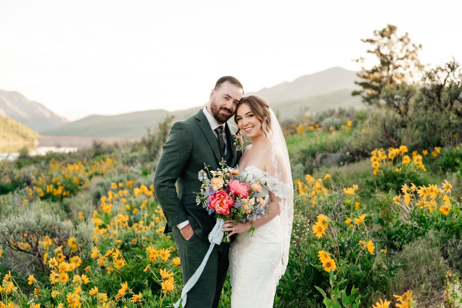 A couple shares an intimate moment surrounded by vibrant yellow wildflowers in a mountain landscape at sunset.