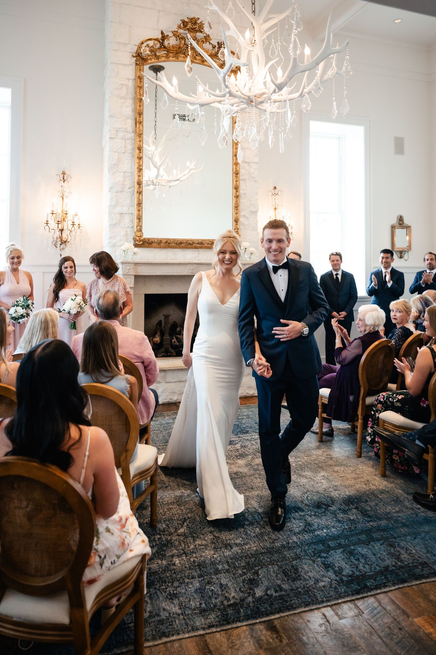 Newlyweds walk down the aisle under a stunning white antler chandelier in an elegant venue.