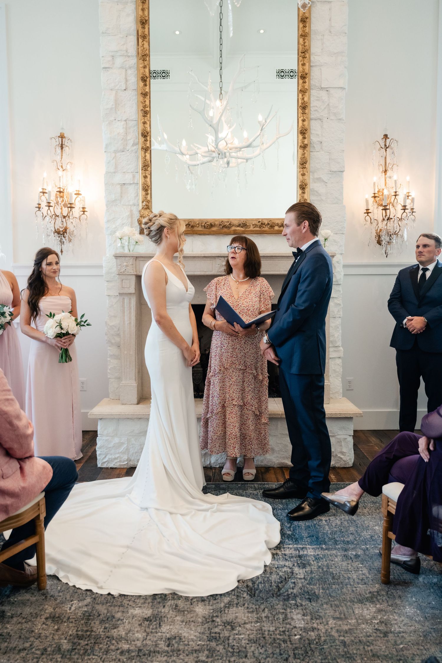 An elegant wedding ceremony in a white room with crystal chandeliers and a long-trained bridal gown.