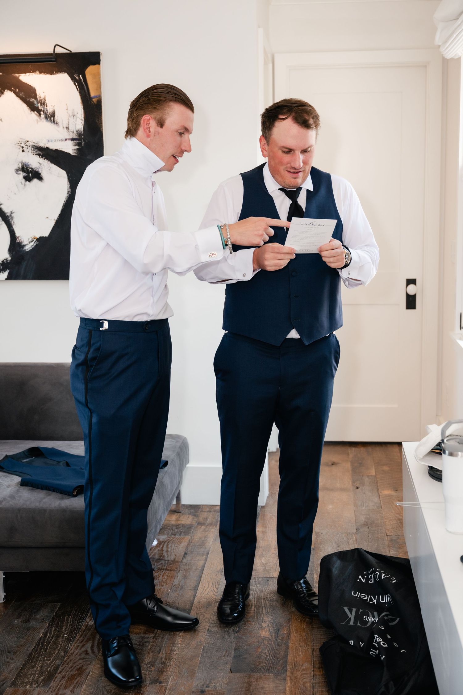 Two men in formal attire looking at a phone in a modern room with abstract art.