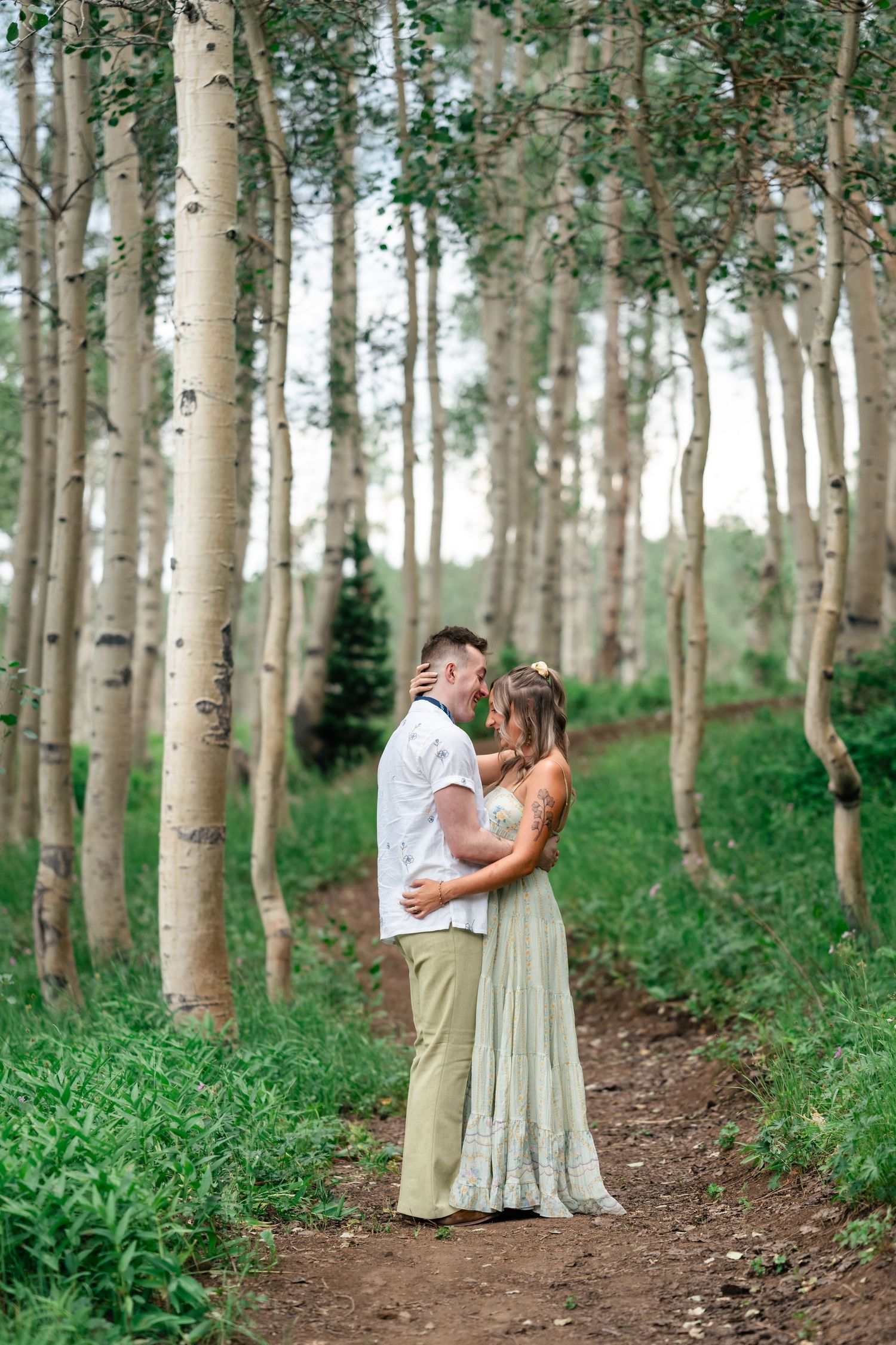A romantic couple embraces on a forest trail surrounded by tall aspen trees and lush greenery. Utah mountain engagement session.