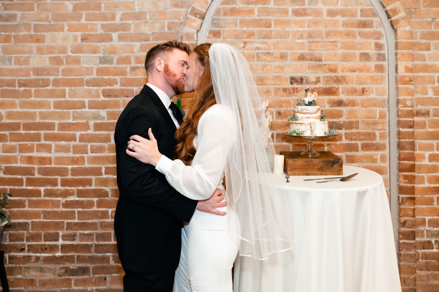 Wedding couple embraces beside rustic wedding cake on table against brick wall at indoor venue.