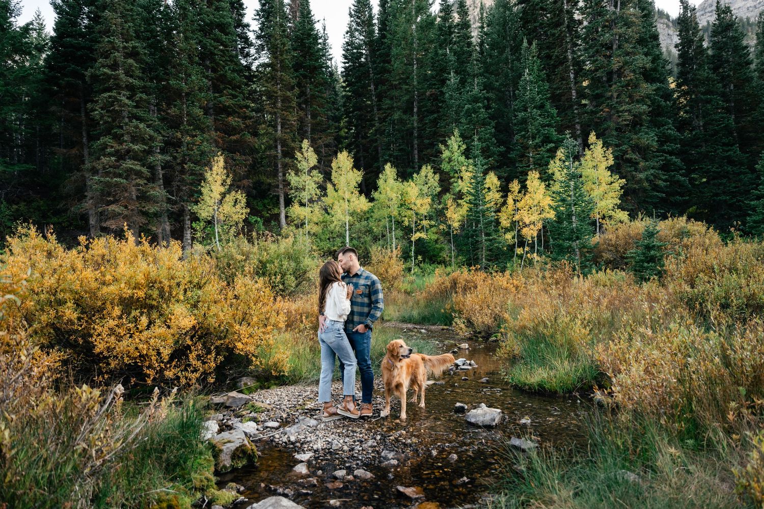 A couple and their dog stand on a rock overlooking a scenic mountain forest landscape in autumn.