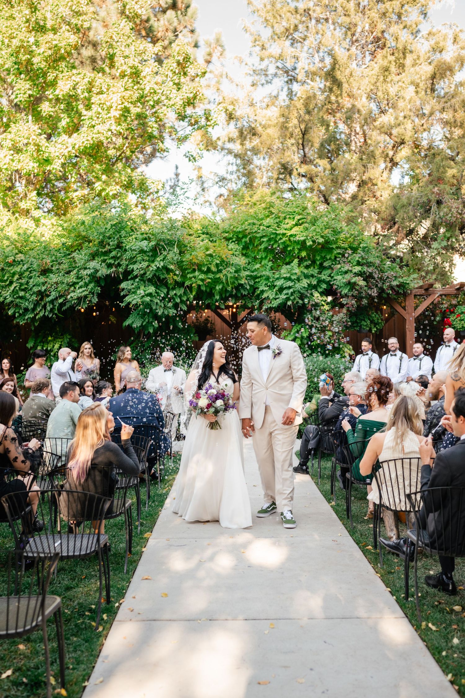A couple walks down an outdoor aisle after their ceremony while guests watch from garden chairs.