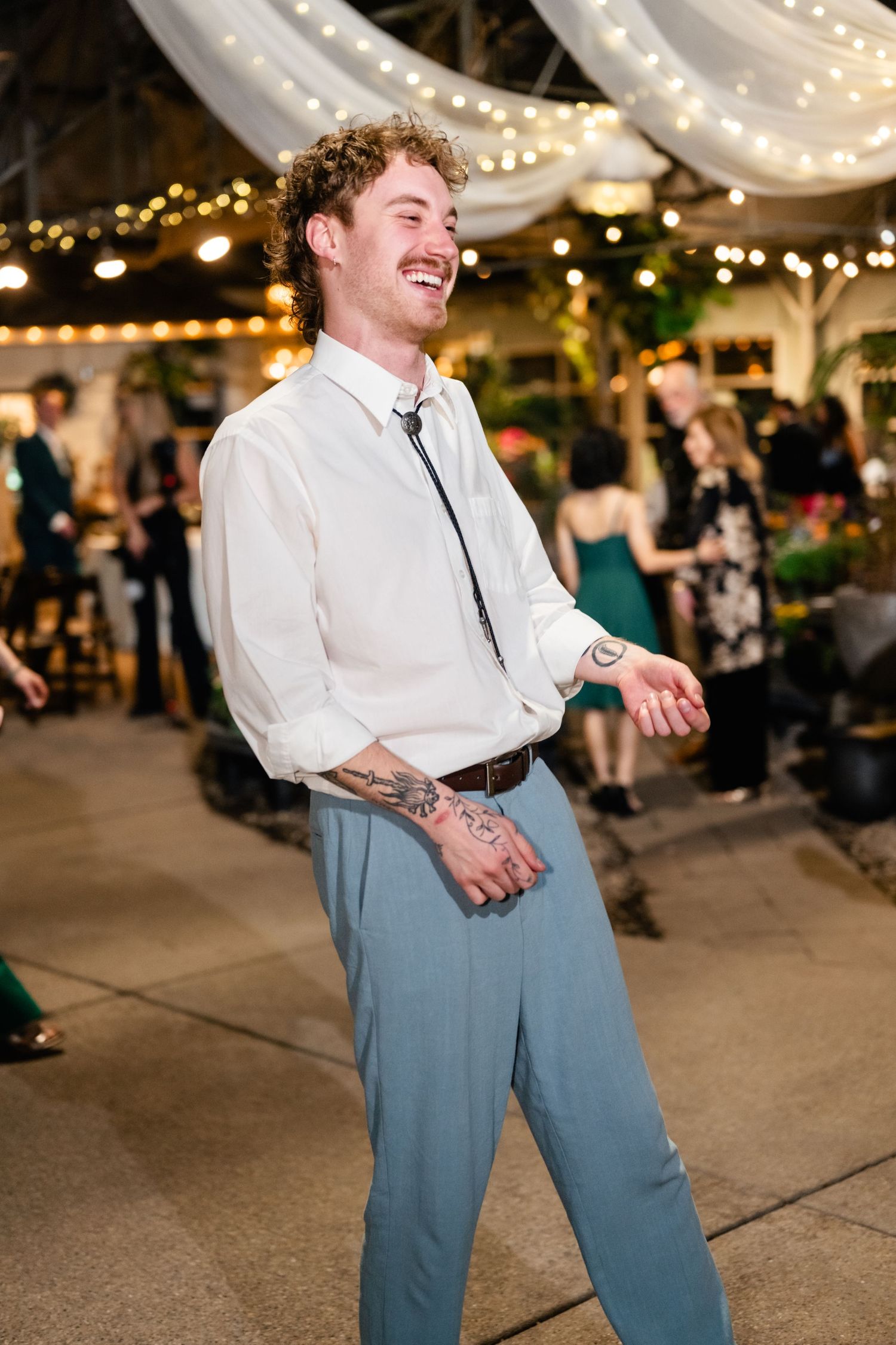 Someone in a white dress shirt and blue pants dances joyfully at an indoor event with string lights and decor.