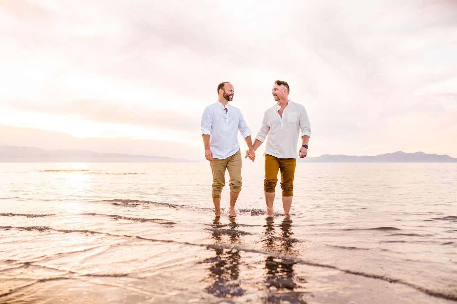 Two people walking hand in hand at sunset on a beach with mountains in the distance.