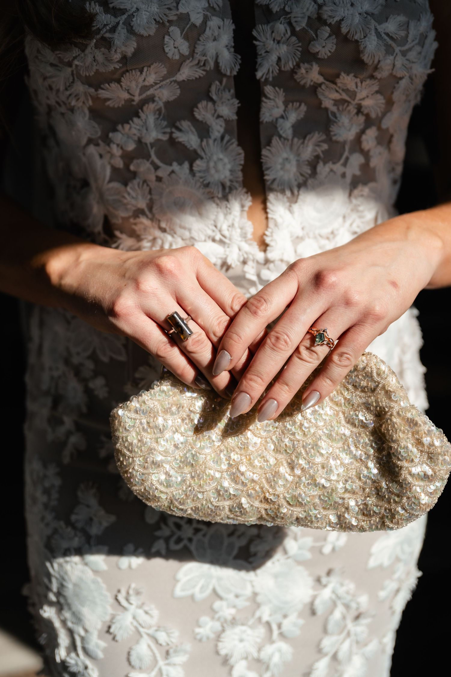 Close up of hands wearing rings holding a sparkly beaded clutch purse against a white lace dress.