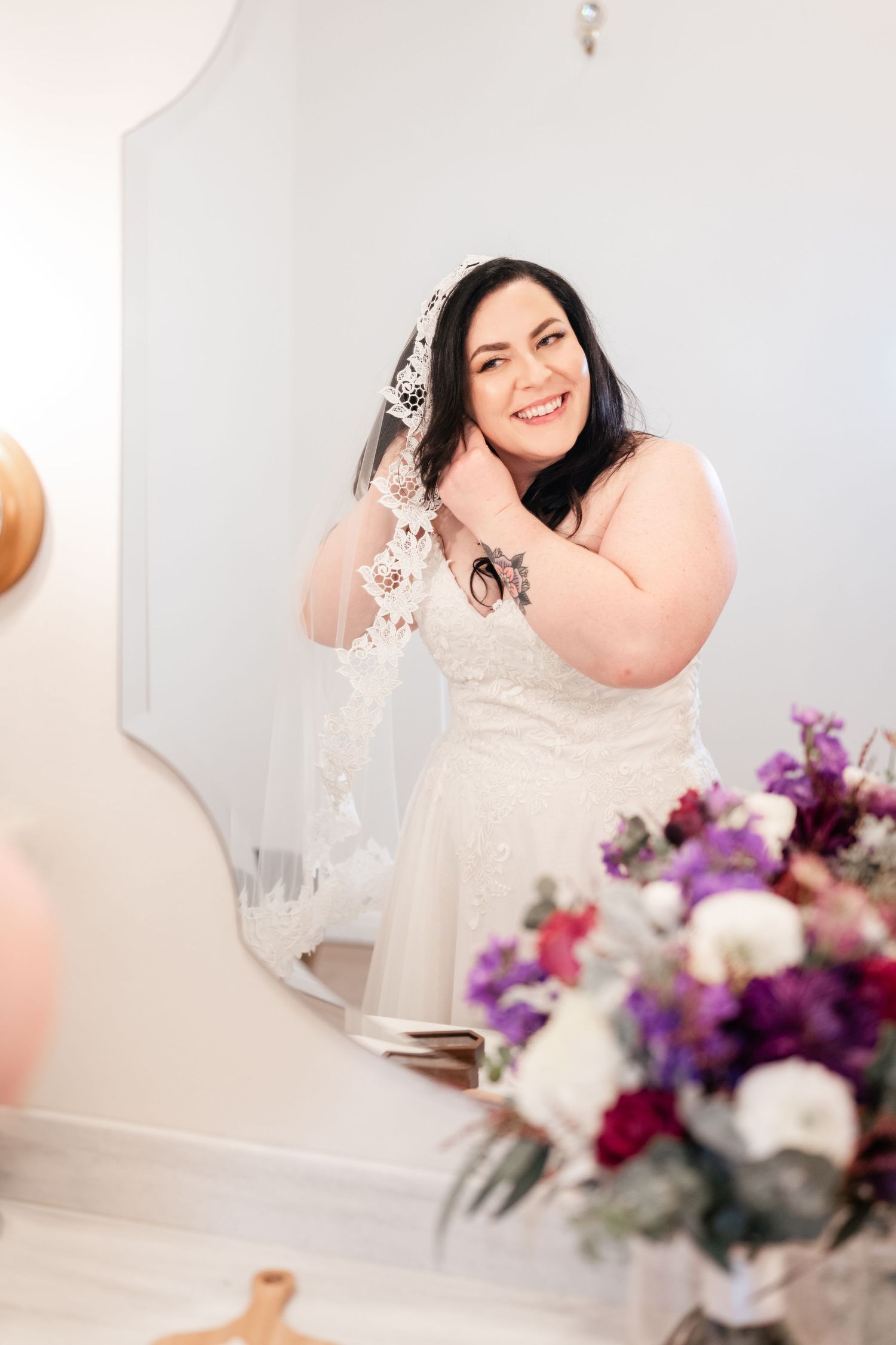 A bride in a white wedding dress with lace veil poses next to vibrant purple and white floral arrangements.