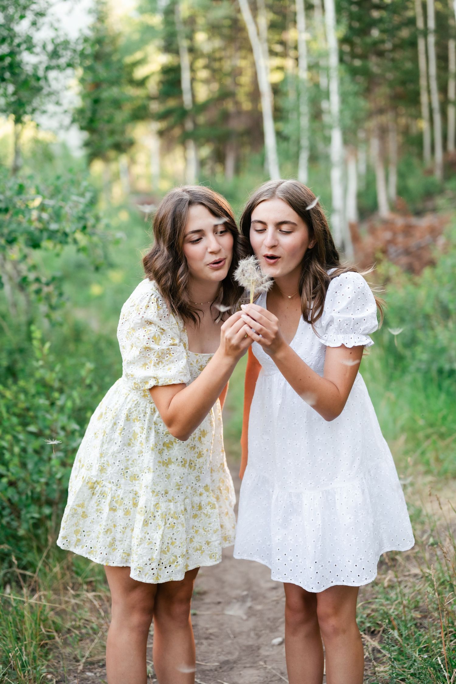 Two people in white dresses share playful moments together while walking through a sunlit forest path.