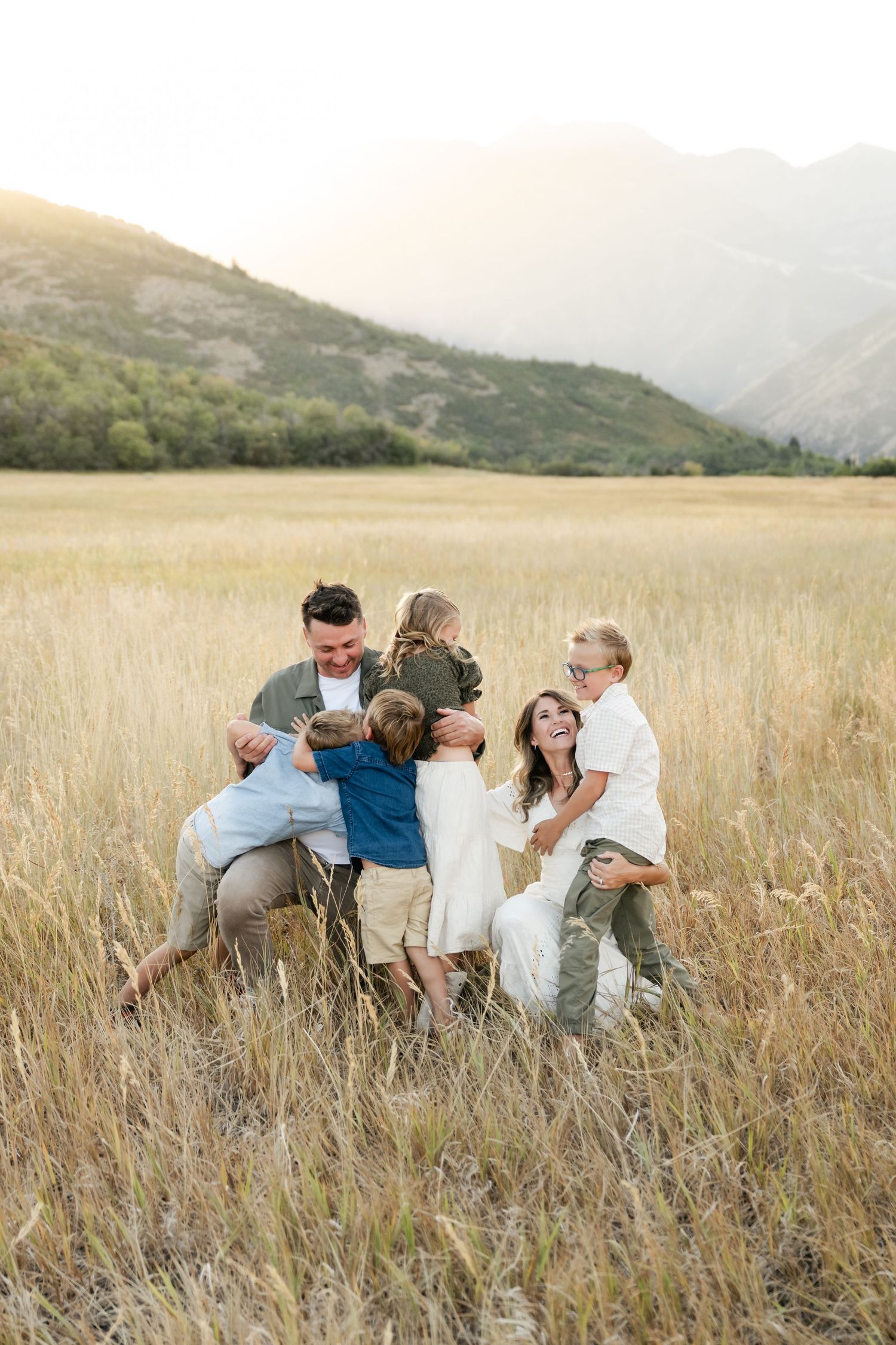 A family sits together in a golden wheat field during sunset with mountains in the background.