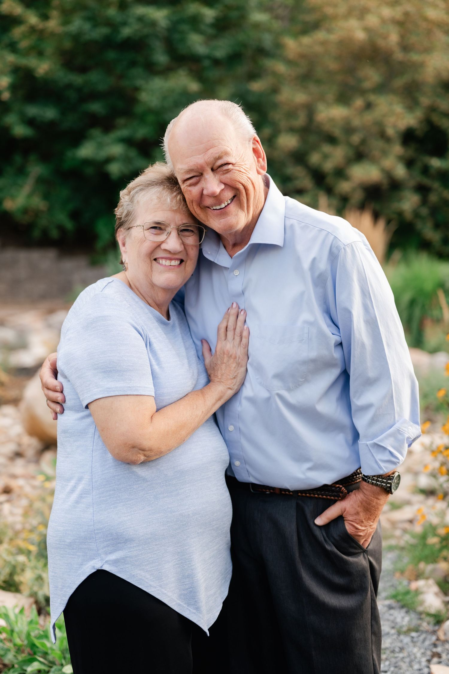 Elderly couple in light blue and dark clothing embracing outdoors while sharing joyful moments together in nature.