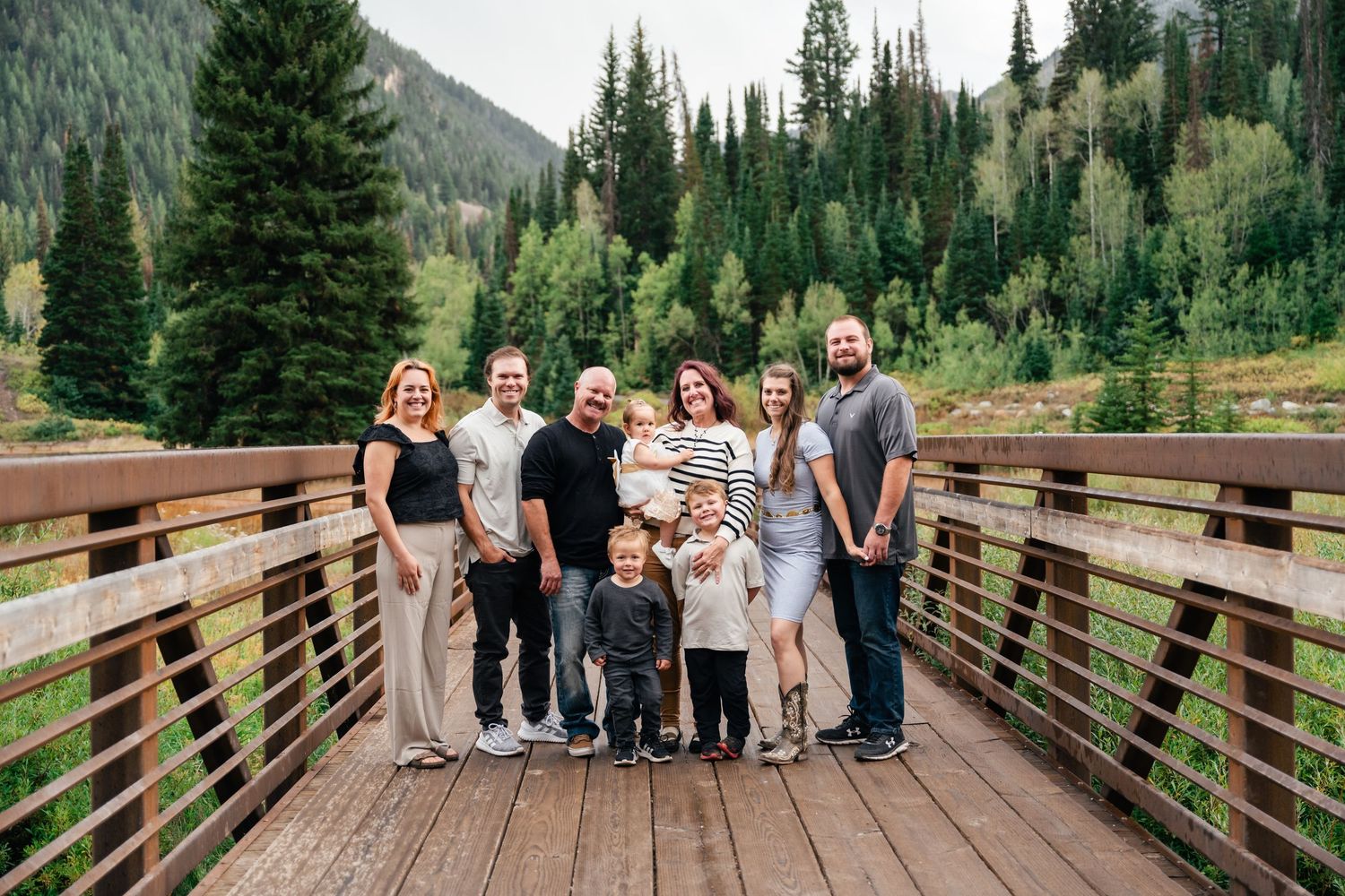 A group of people stand on a wooden bridge surrounded by evergreen trees and mountains in a scenic outdoor setting.