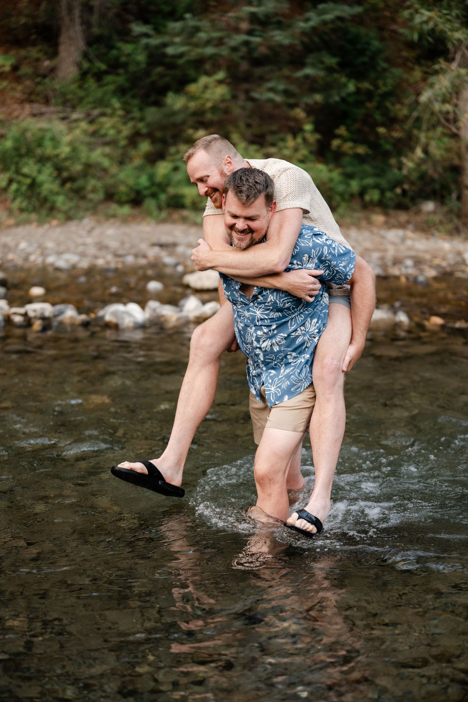 Two men laughing and enjoying a piggyback ride in a shallow rocky river surrounded by lush green trees.