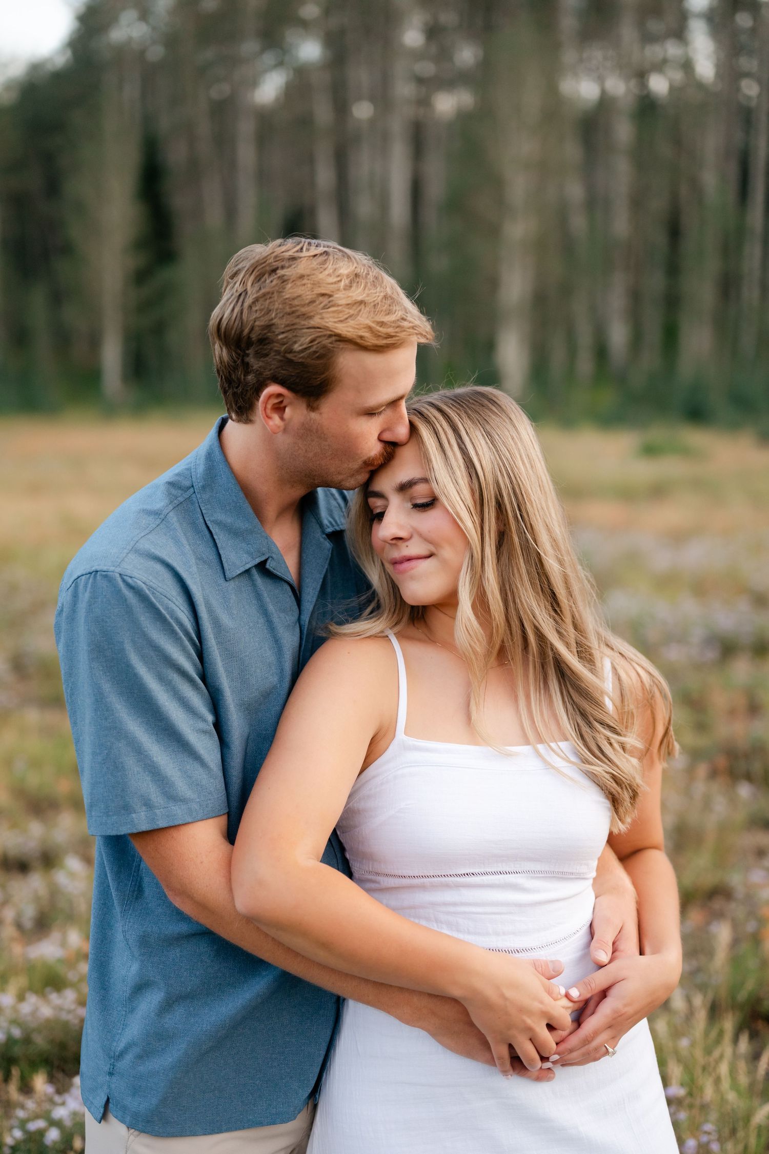 A couple embraces in a field, man kissing woman's temple, trees in background.  Mountain engagement photos near Park City.