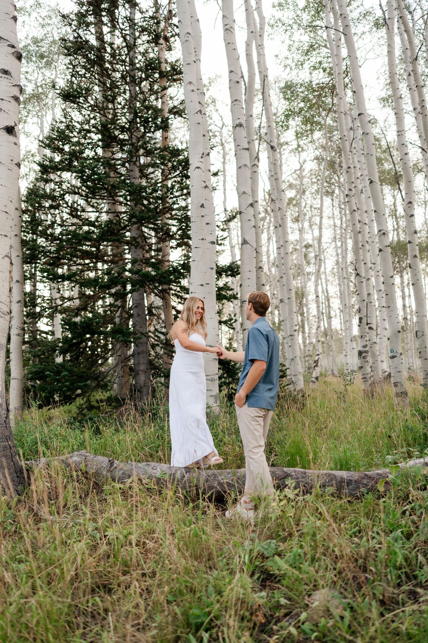 Couple holding hands in an aspen grove, woman in white dress, man in blue shirt, lush green grass.