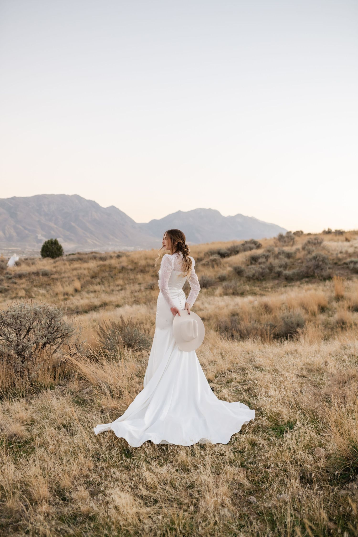 Bride in flowing white dress stands against mountain backdrop in golden field during sunset. Utah mountain elopement photographer.