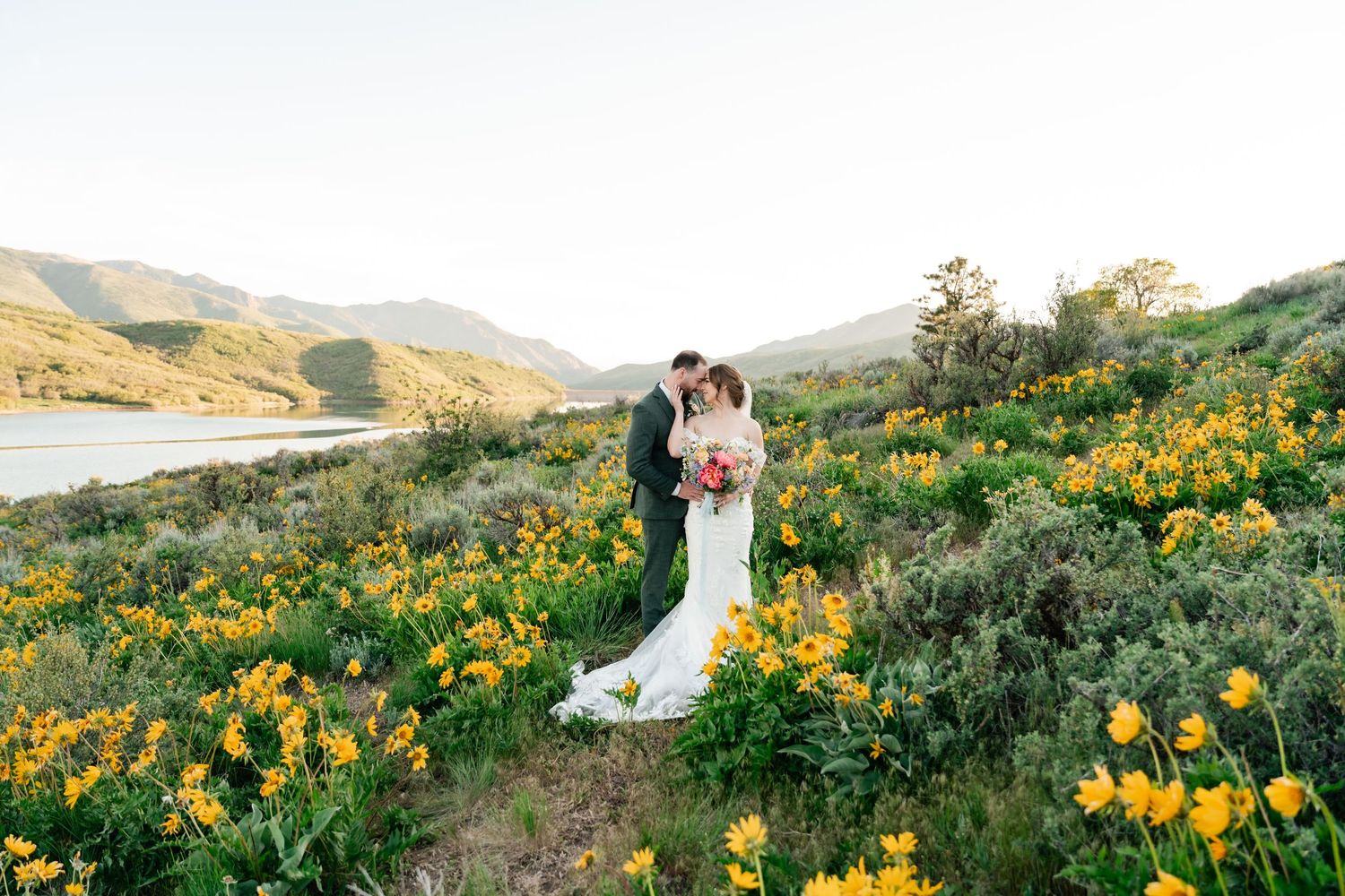 A couple stands among vibrant yellow wildflowers on a mountainside during golden hour with rolling hills in the background. Utah Weddings.