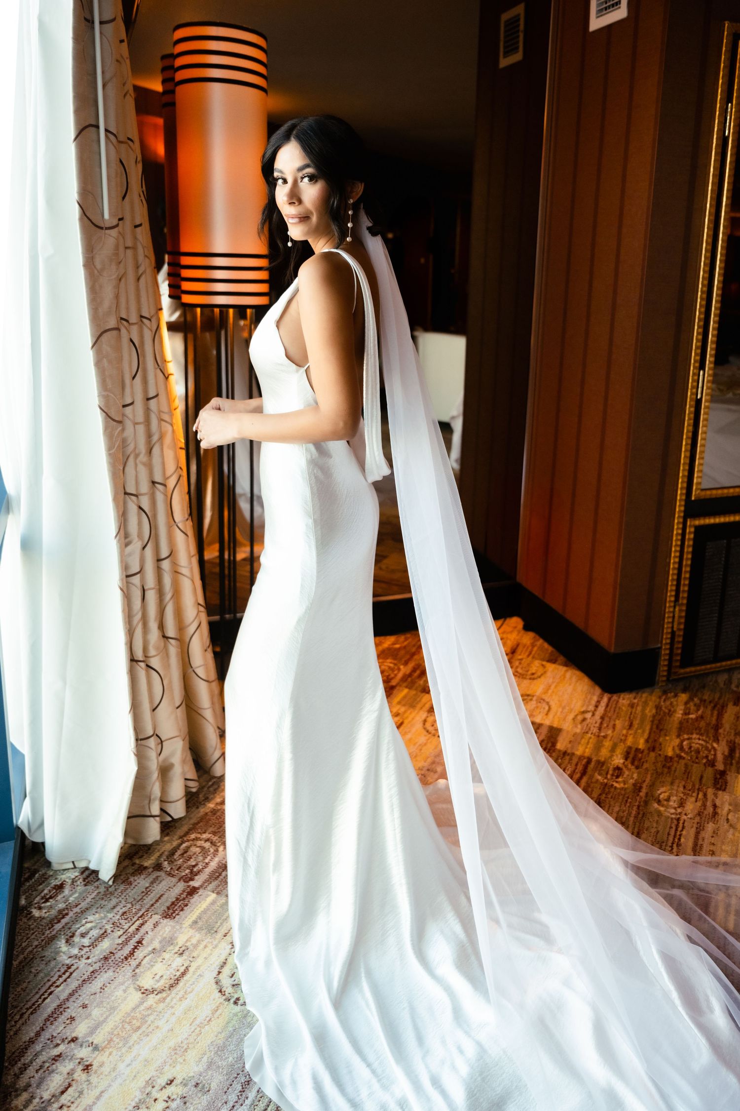 A white wedding dress being adjusted and styled in an elegant hotel room with warm lighting.