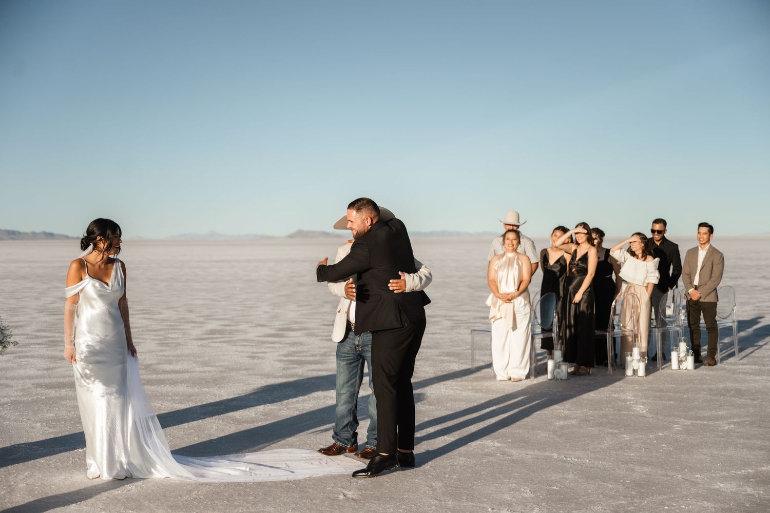 Wedding party and guests gathered on salt flats with long shadows cast on white ground.