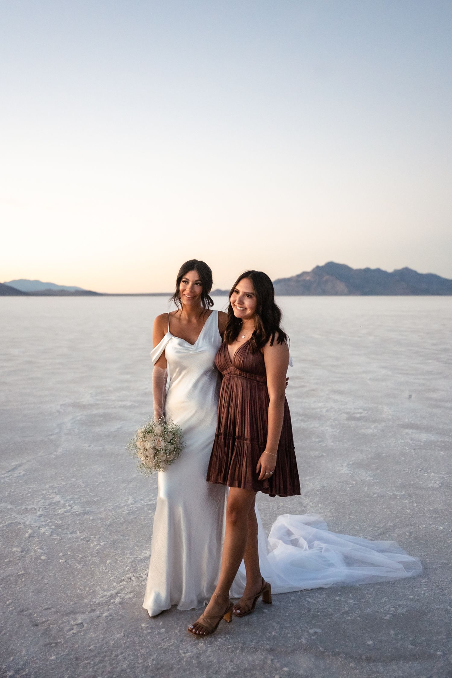 Two people in contrasting outfits embrace while standing on salt flats with mountains in background.