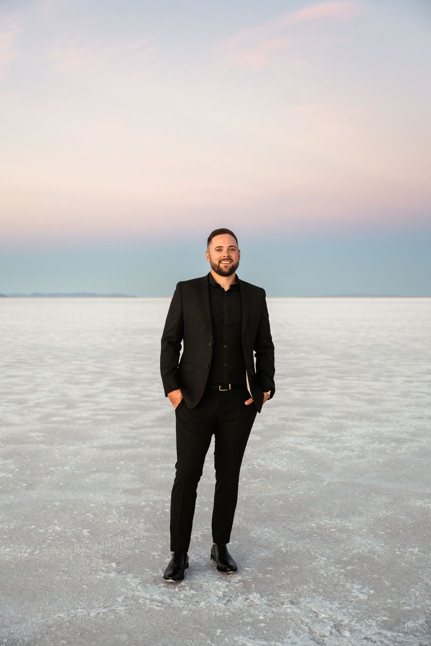 Person in black suit poses on expansive salt flats during sunset.