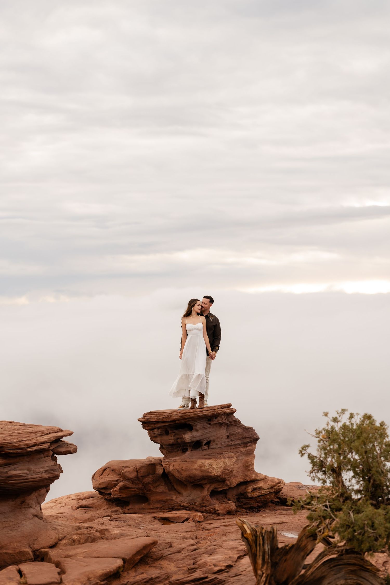 A couple stands embracing on red rock formations during sunset with cloudy skies in the background.