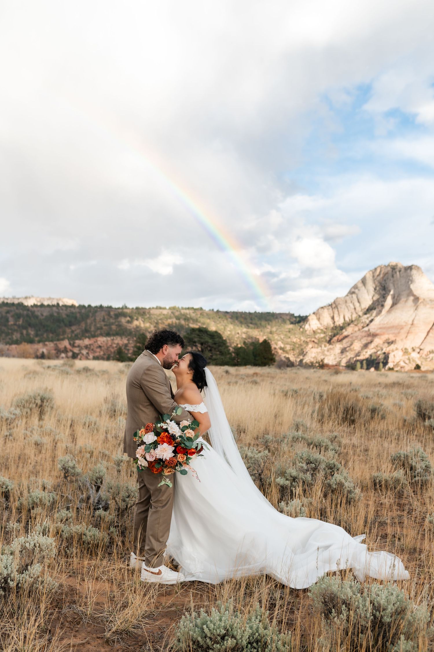 A romantic wedding couple embraces in a golden field with dramatic mountain cliffs and blue skies in the background.